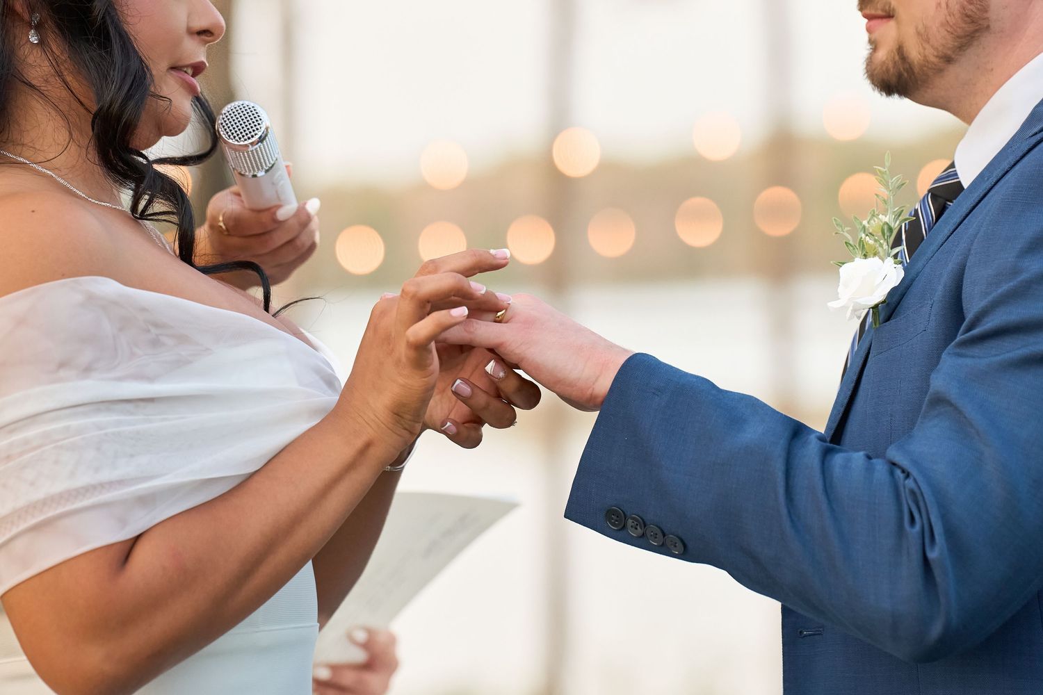 Bride placing wedding ring on groom's finger during outdoor ceremony, both dressed in elegant wedding attire.