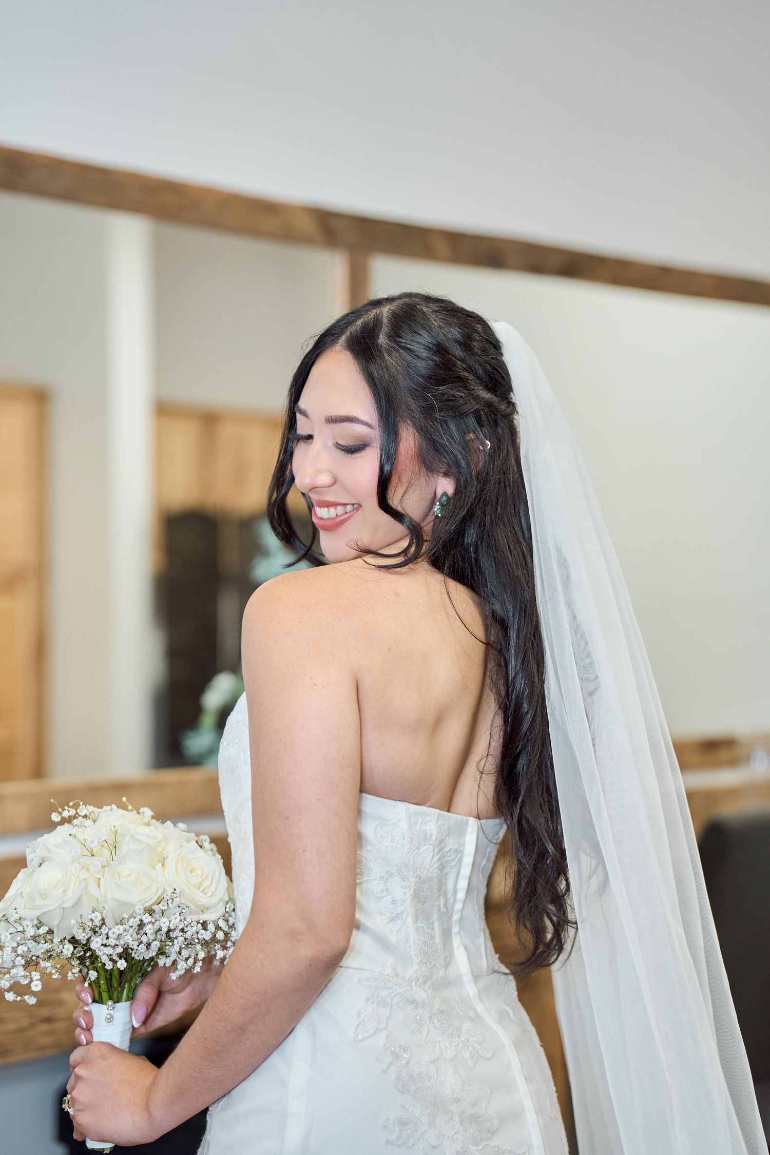 A bride in a strapless white wedding gown holds a baby's breath bouquet while looking down with a flowing veil.