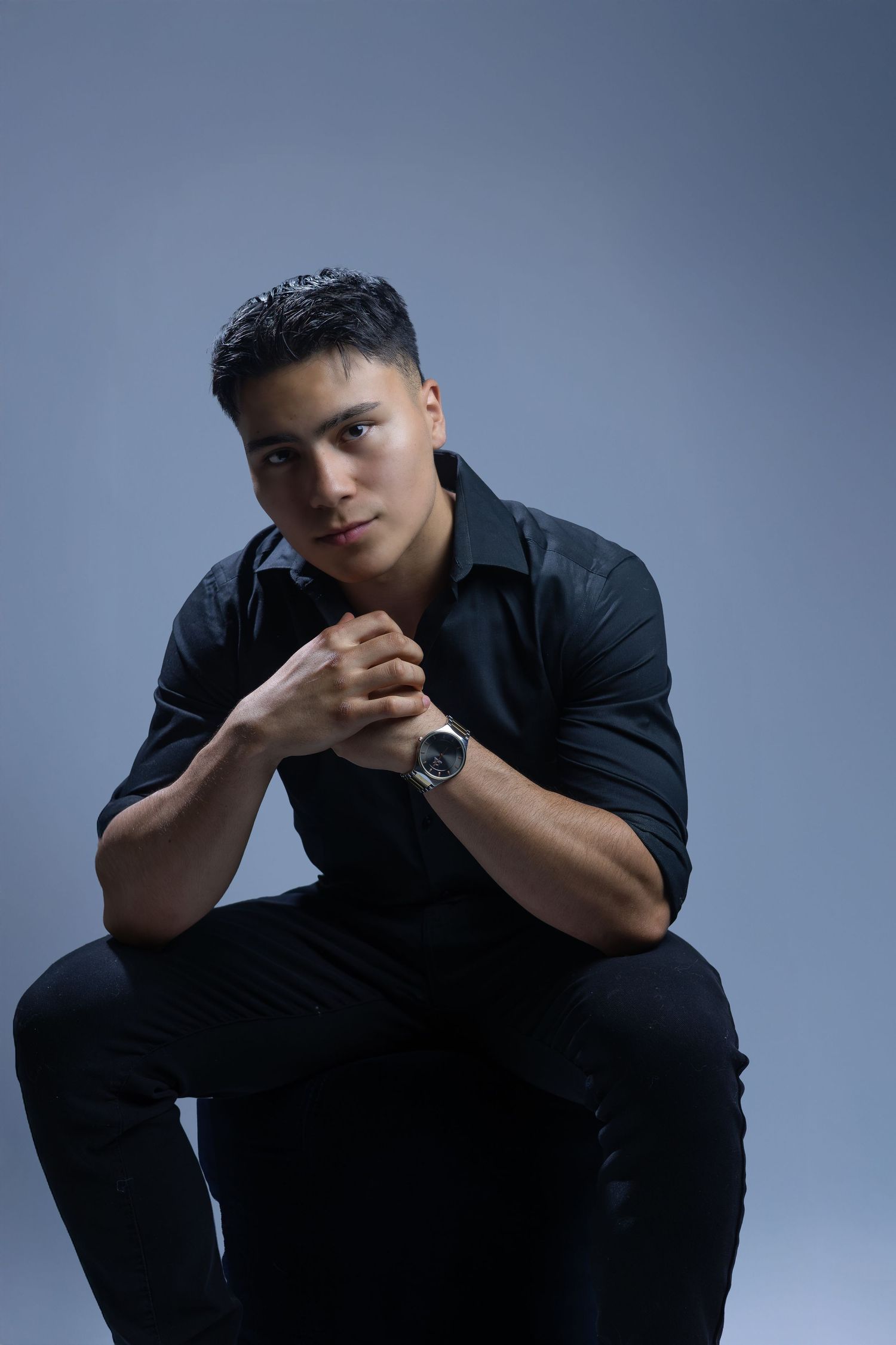 A person in a black shirt sits pensively with clasped hands against a blue-gray backdrop, wearing a sleek watch.