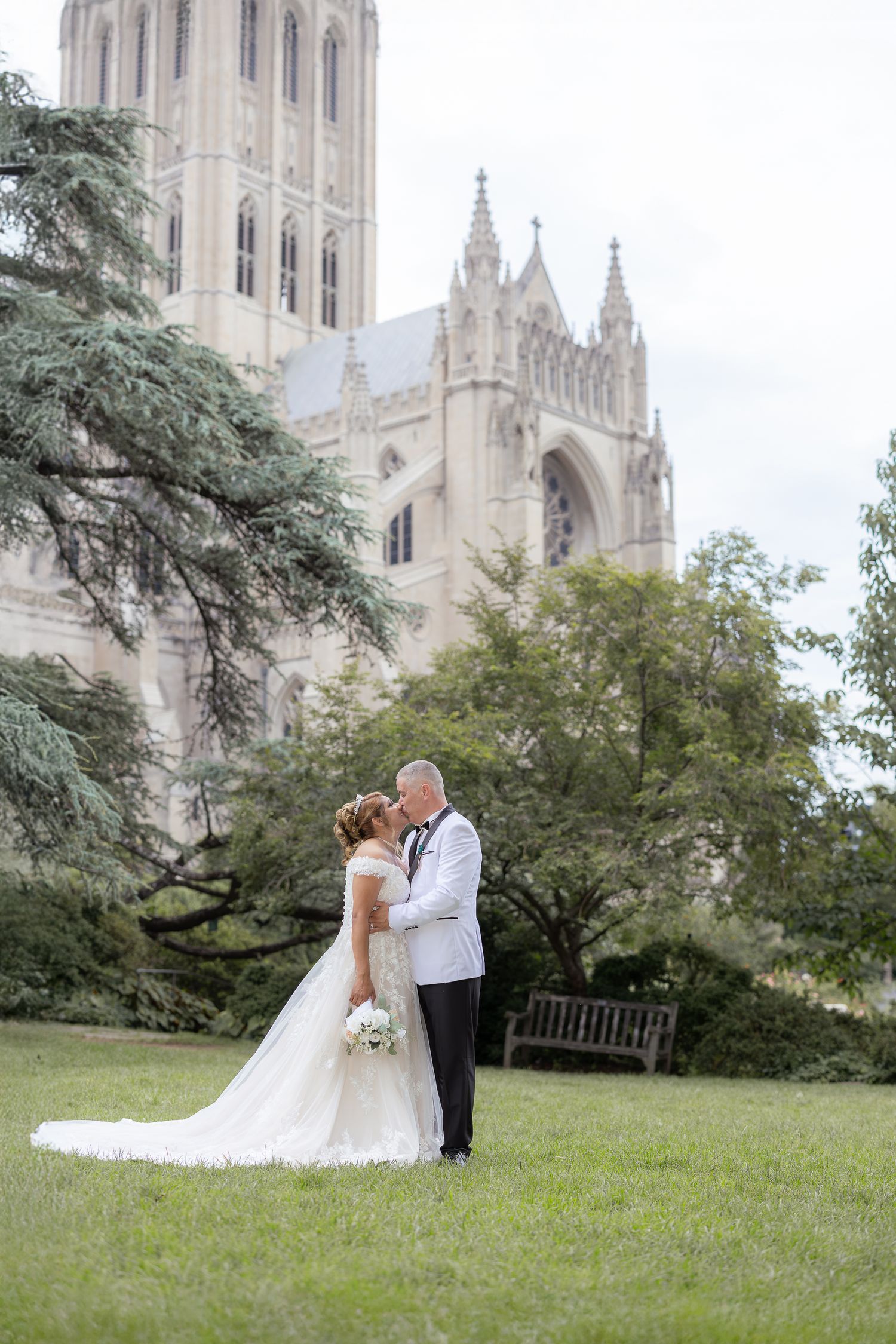 A couple shares an intimate moment in their wedding attire on a grassy lawn in front of a majestic gothic cathedral.