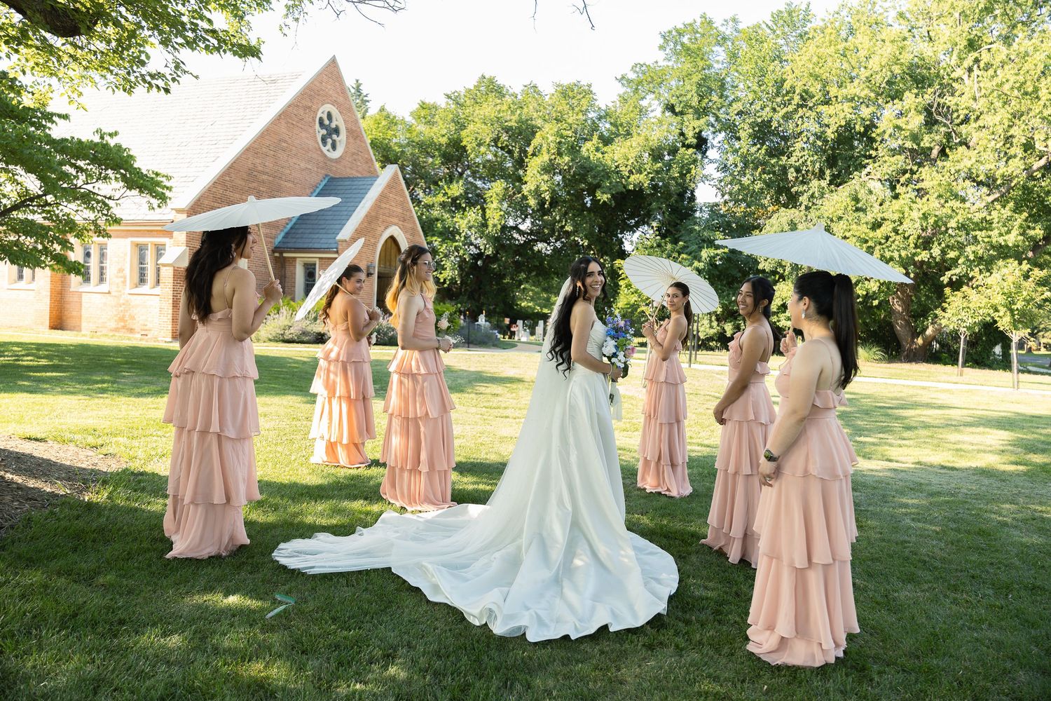 A wedding party gathers on a sunny lawn outside a charming chapel with bridesmaids in blush pink dresses holding parasols.