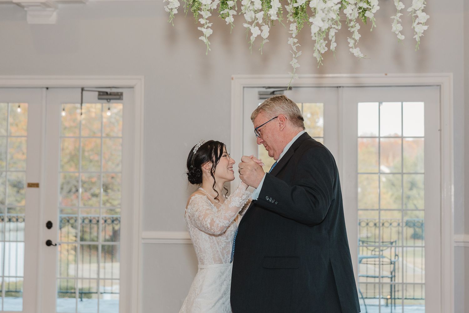 Elegant white floral arrangements hang from the ceiling in a bright reception venue with large windows.