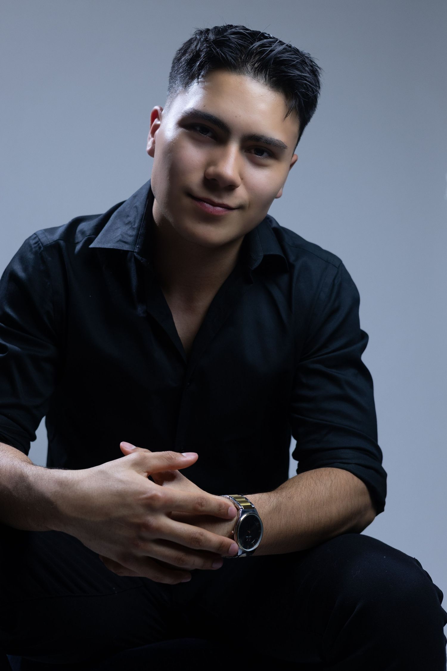 A person in a black button-down shirt sits with clasped hands and wears a watch against a gray background.