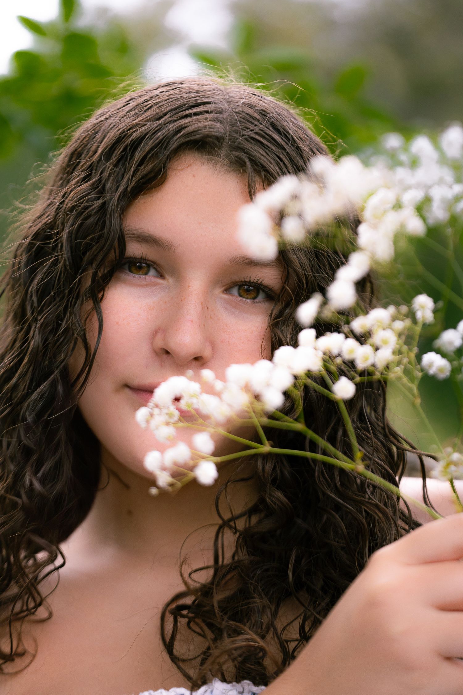 Dark-haired woman with curly hair posing in nature with delicate white flowers in soft outdoor lighting.