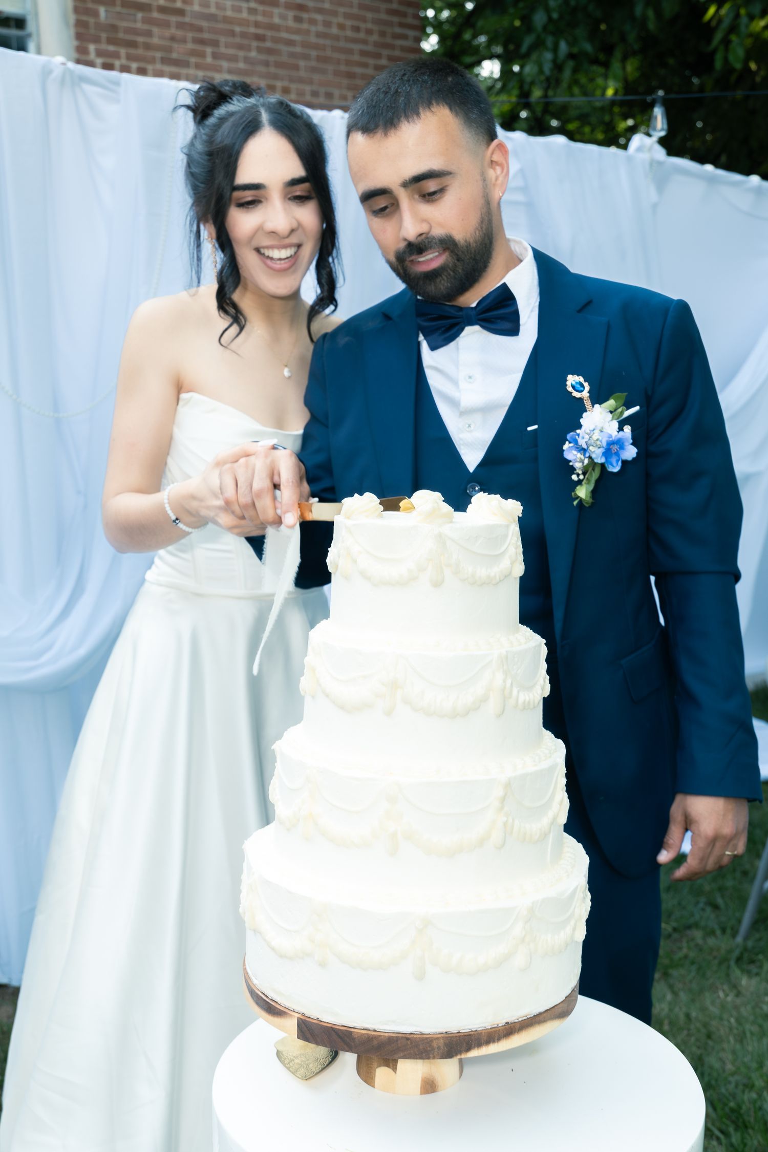 Newlywed couple cutting into their elegant white four-tier wedding cake with decorative swirls and gold accents.