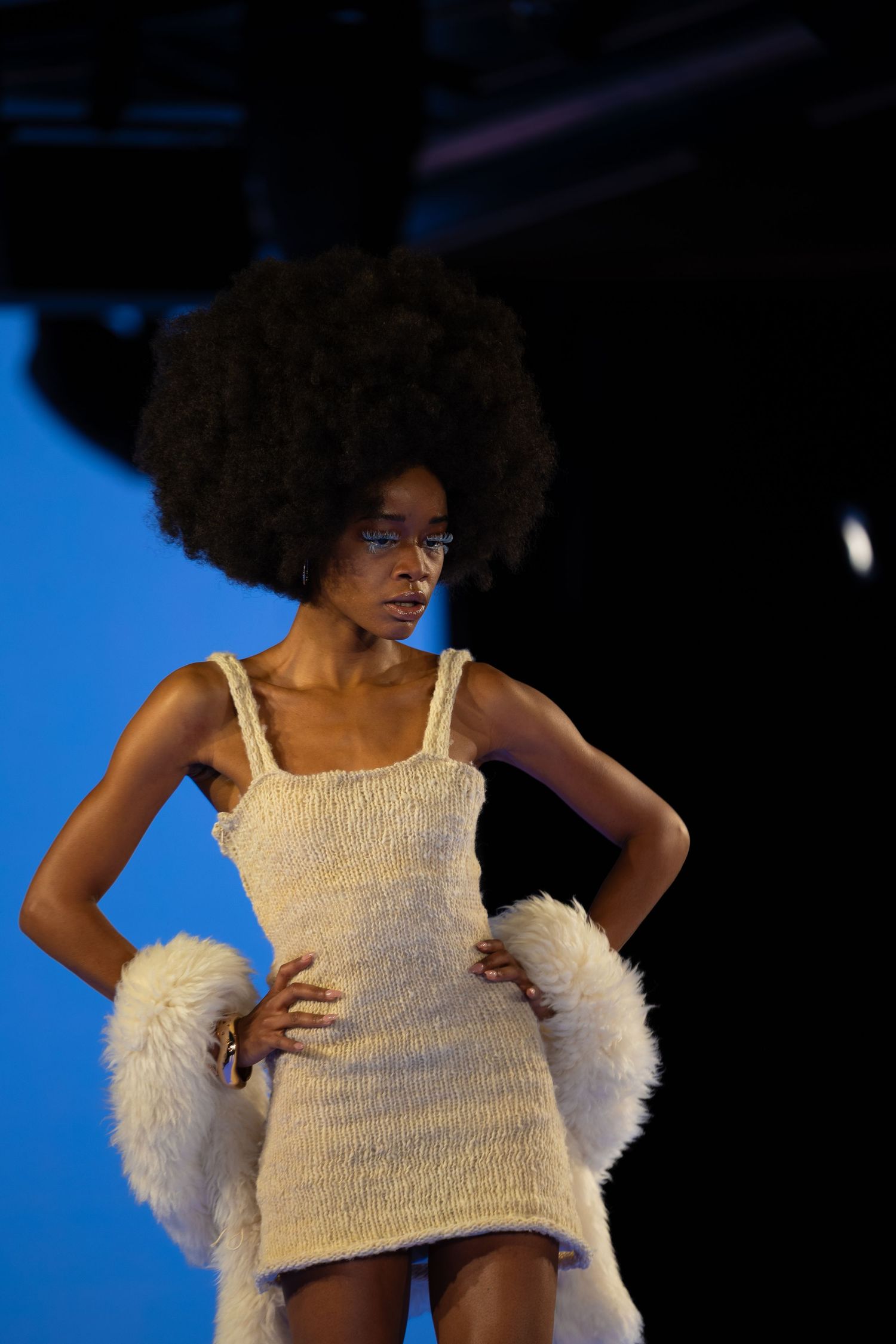 Model in cream knit dress with fur accessories poses against blue backdrop on runway.