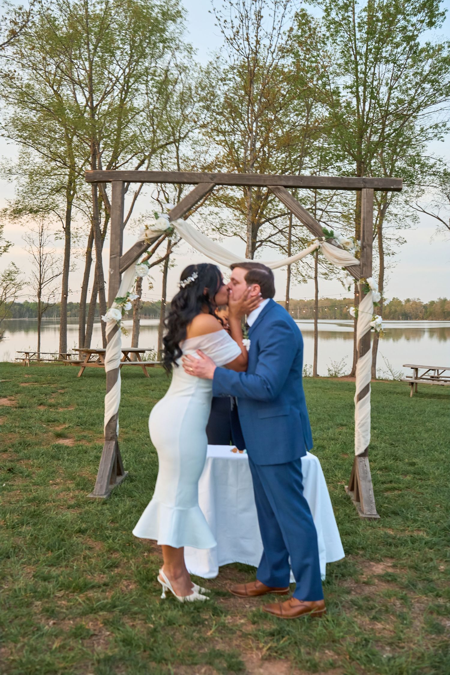 Bride and groom share first kiss under rustic wooden arch by lakeside at outdoor wedding ceremony.
