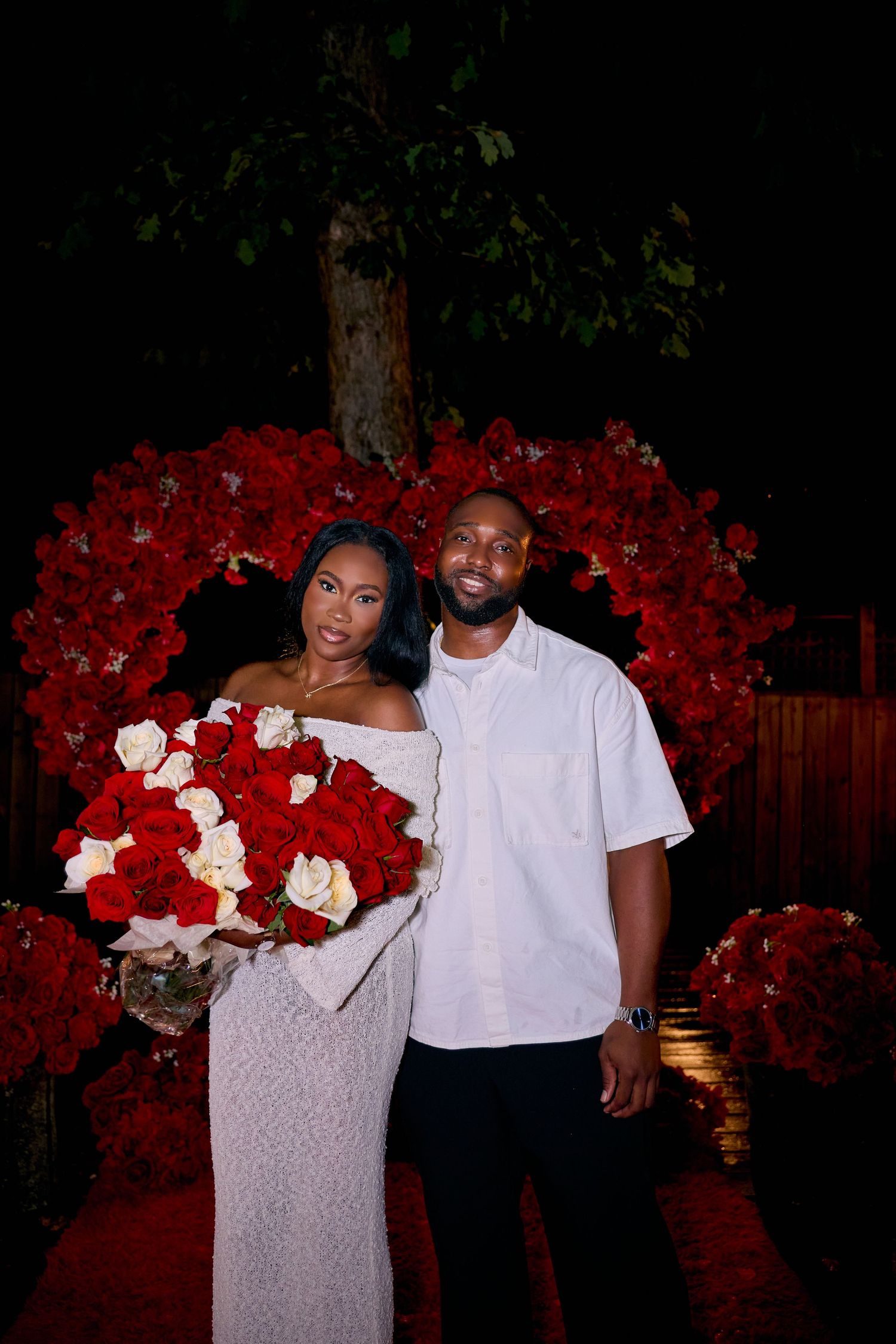 A couple poses in front of a romantic red rose arch during an evening photoshoot.