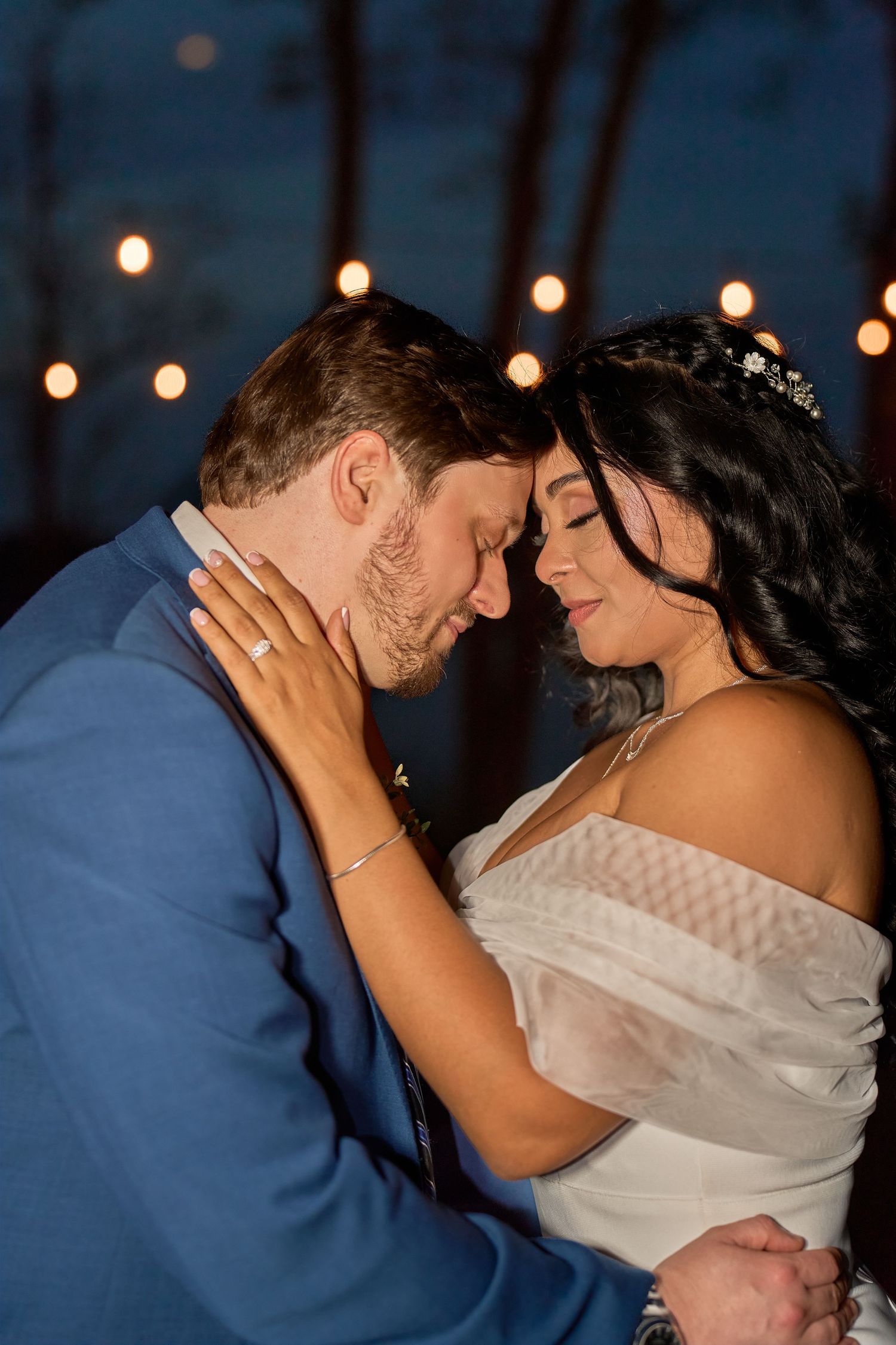 Bride and groom share an intimate moment under string lights at night, with bride in white off-shoulder gown and groom in blue suit.