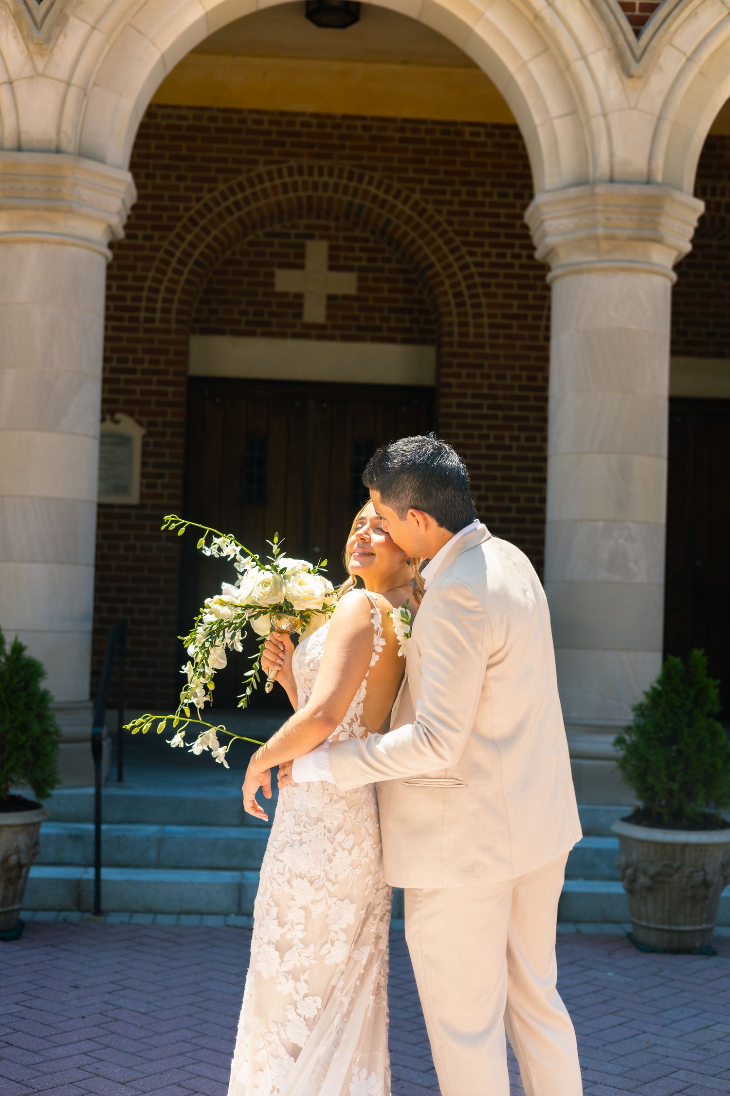 A couple in white attire embraces under an ornate stone archway with brick walls and columns.