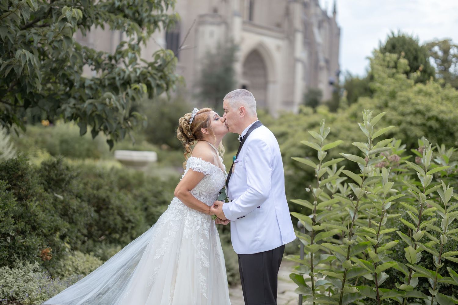 Couple shares romantic wedding day kiss in garden setting with elegant church backdrop.