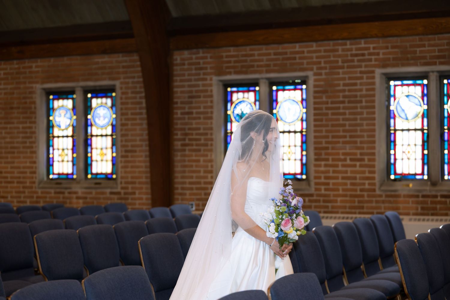 A bride in a flowing white dress stands in a church sanctuary with stained glass windows and wooden pews.