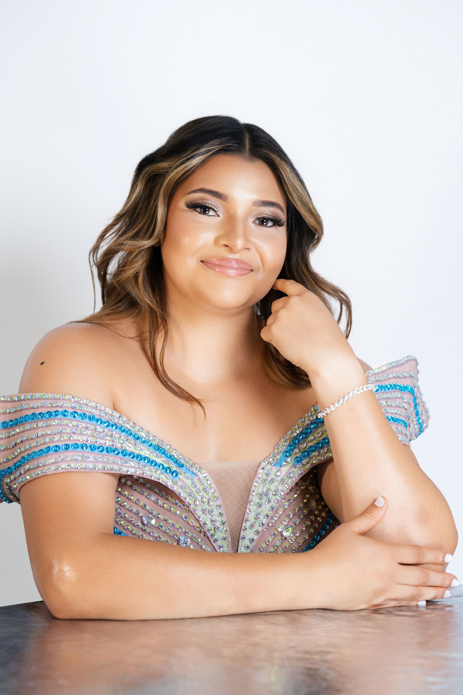 Young woman in a beaded off-shoulder gown with blue accents, smiling gracefully at a table.