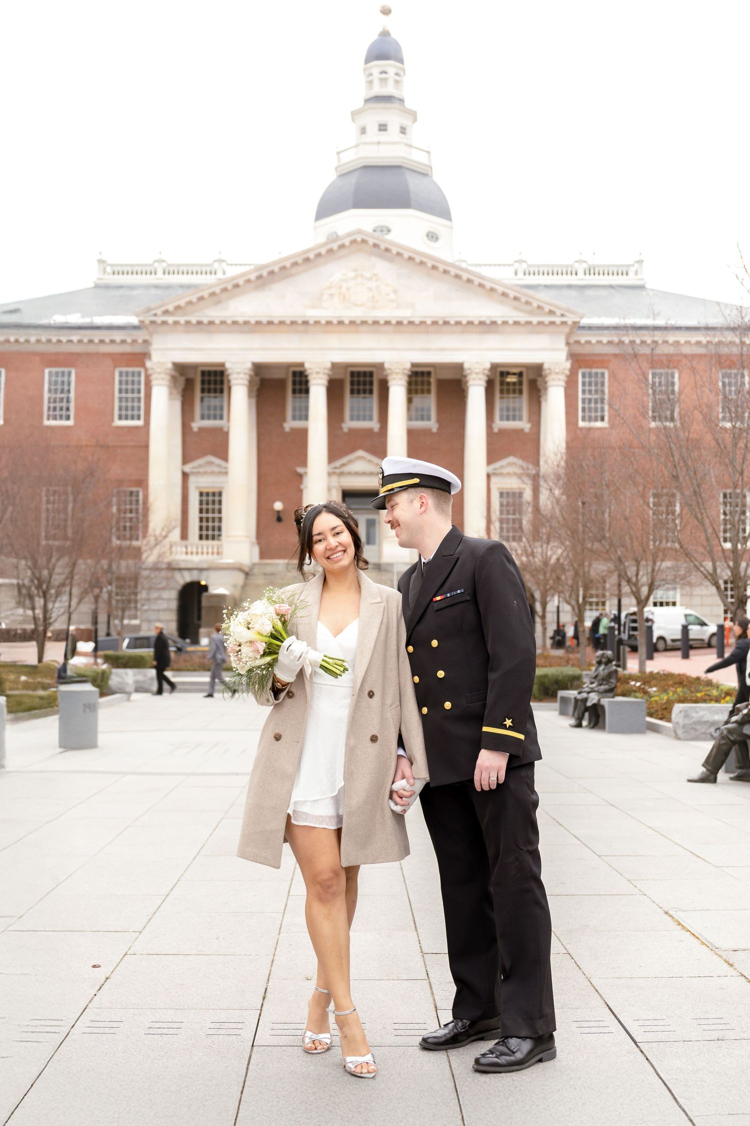 Wedding couple poses in front of historic brick building with white columns and dome architecture.