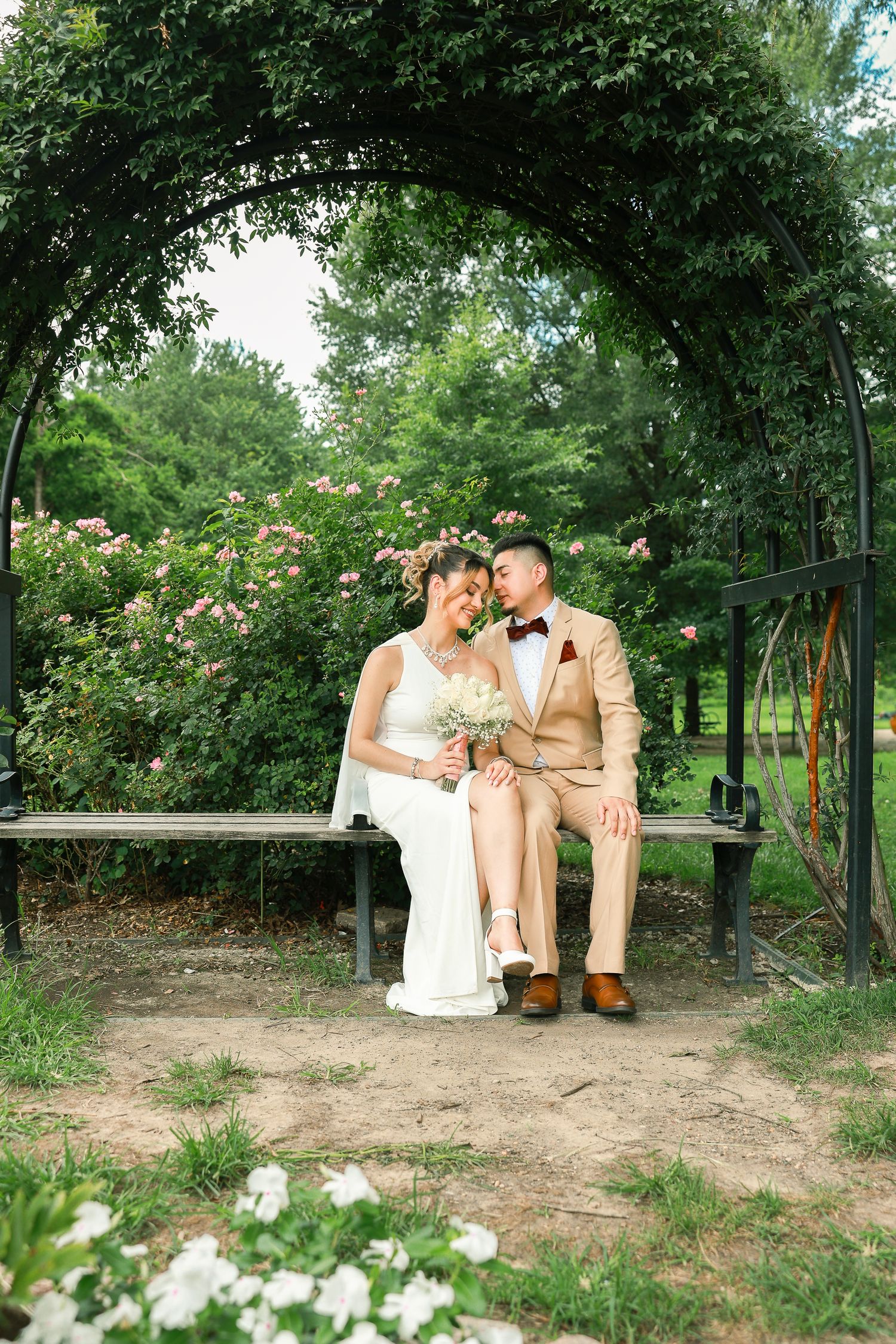 A romantic wedding photo under a lush garden arch with pink roses as a couple sits on a wooden bench.