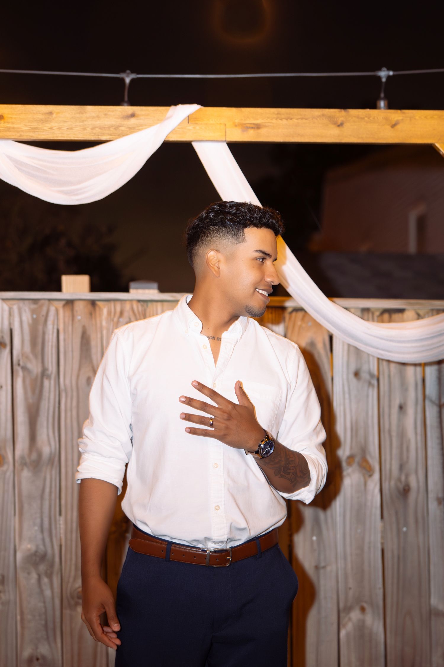 Person in white button-down shirt and navy pants poses against rustic wooden wall with draped white fabric decor.