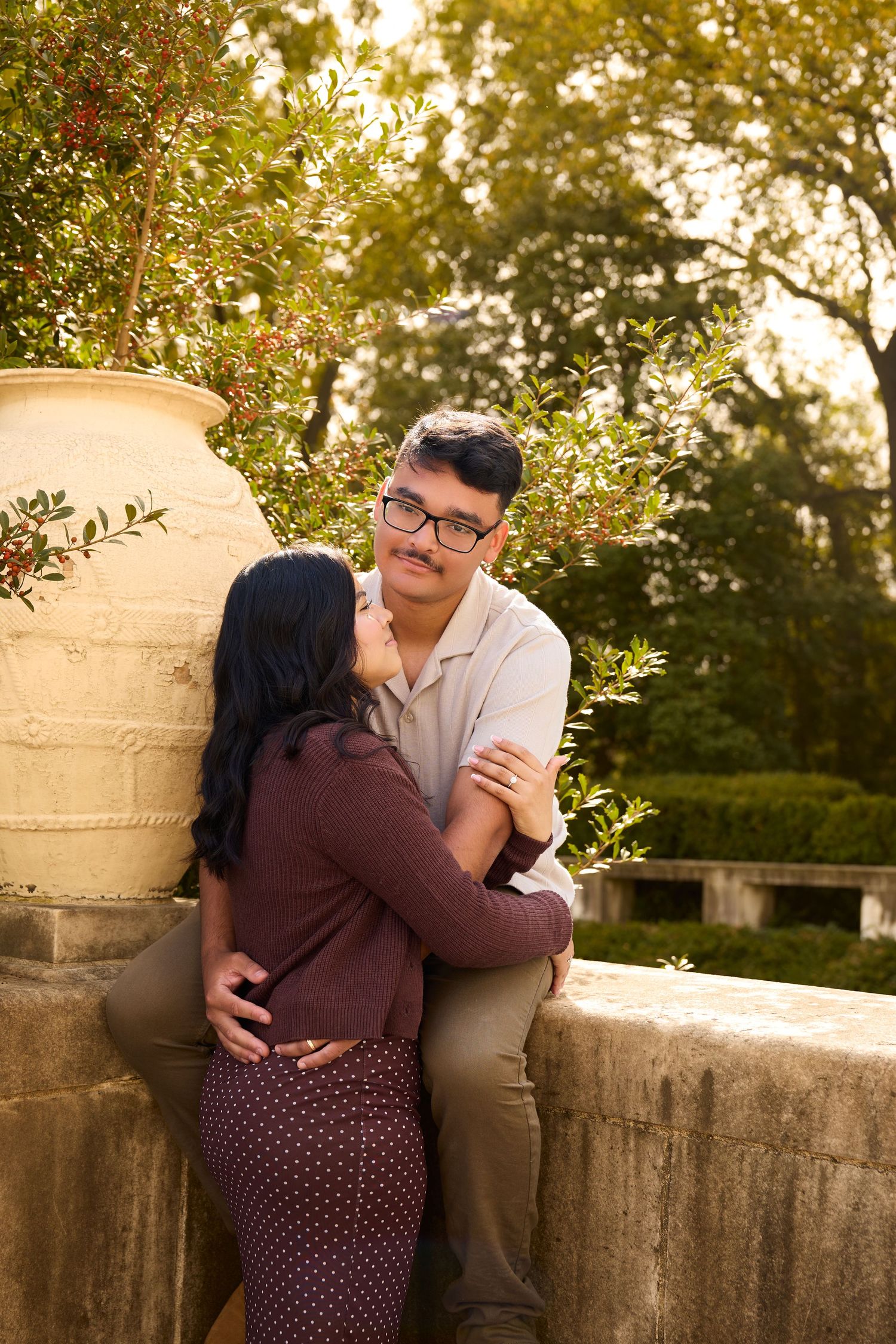 A romantic couple embraces on a stone balcony at sunset with autumn foliage in the background.