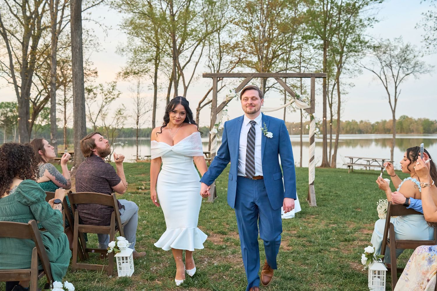 Bride and groom walk down outdoor aisle at lakeside wedding ceremony, guests seated on wooden chairs.