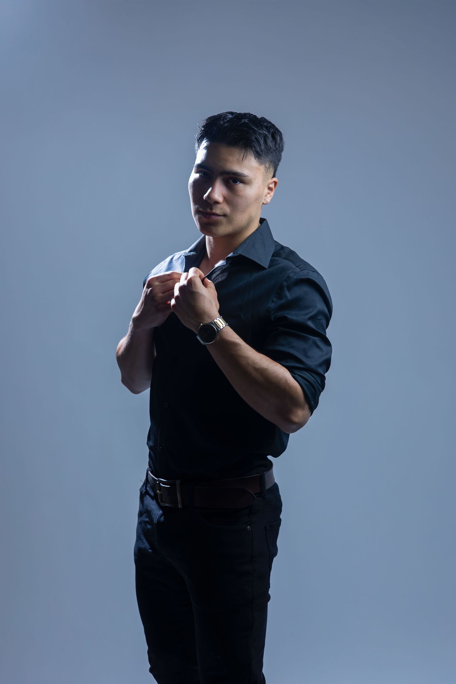 Figure in a dark button-up shirt posing confidently against a gray backdrop with dramatic lighting.