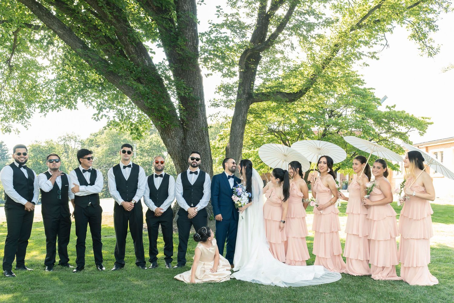 Wedding party in coordinated attire poses in a lush garden setting under green trees.