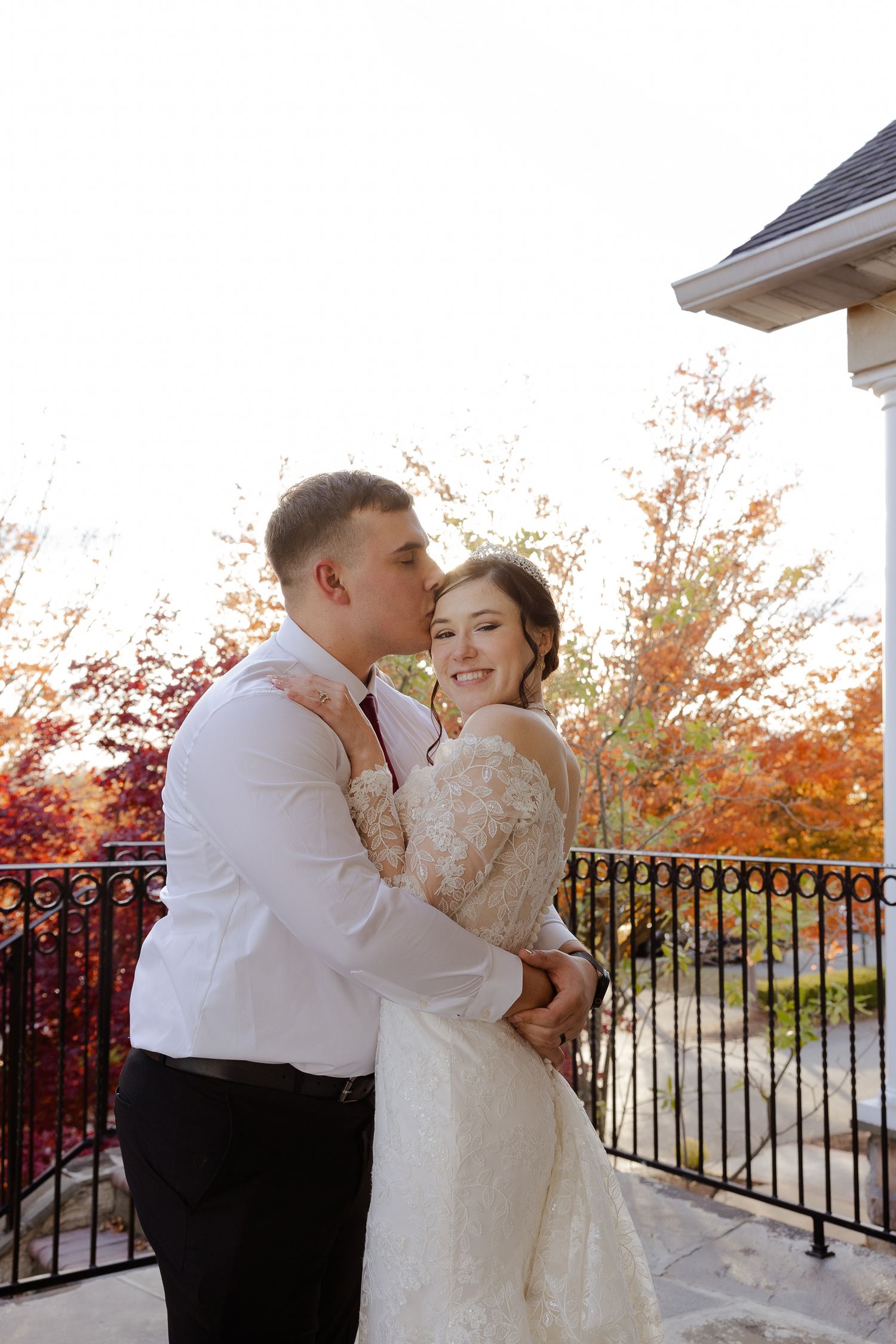 A couple shares romantic moments on a balcony with autumn foliage in the background during sunset.