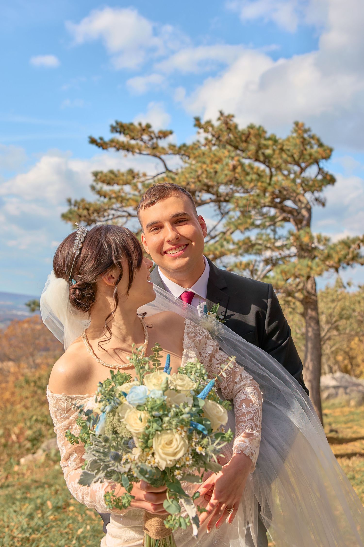 A romantic autumn wedding photoshoot outdoors with a couple embracing under pine trees and blue skies.