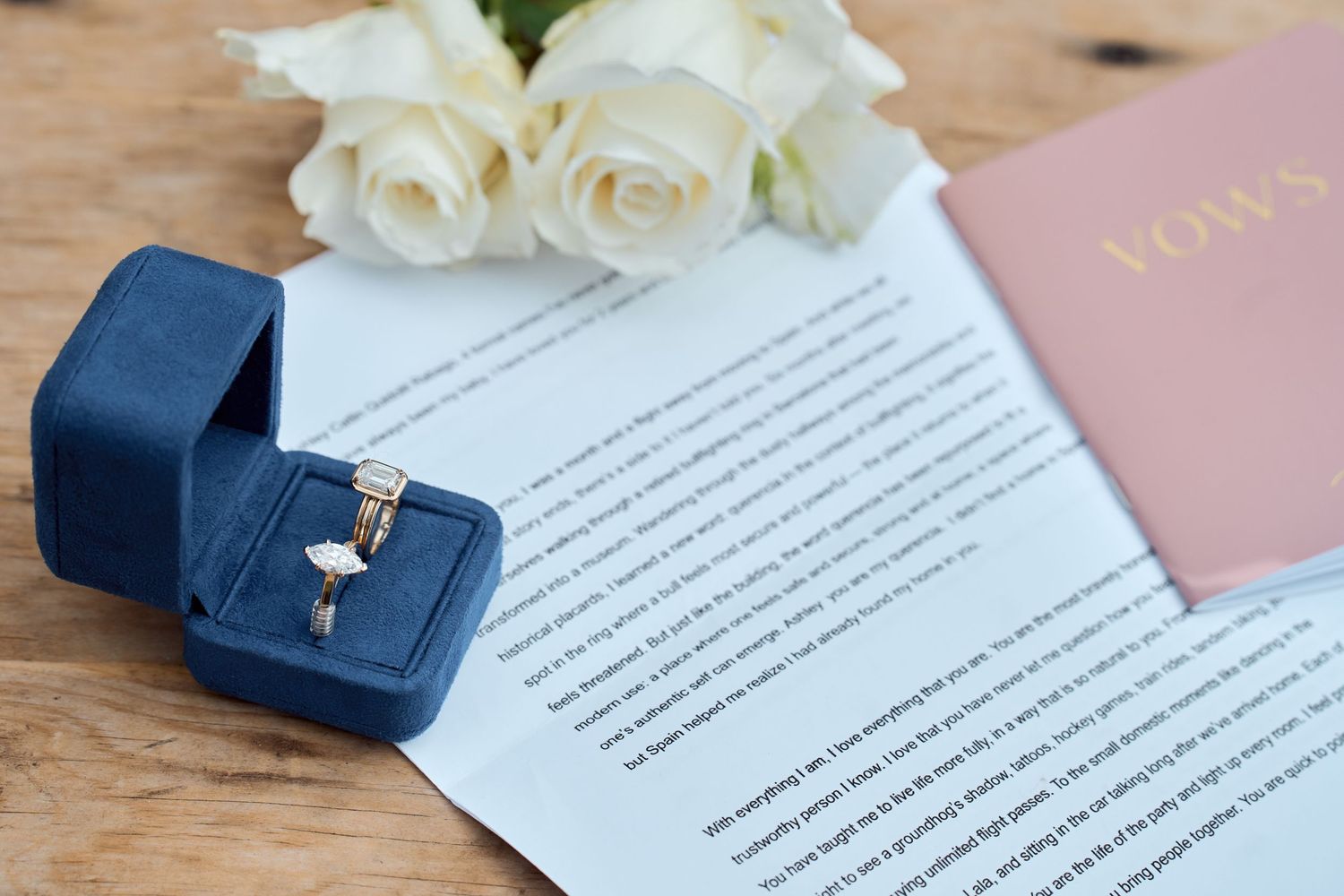 White roses next to wedding documents and navy blue ring boxes on wooden surface.