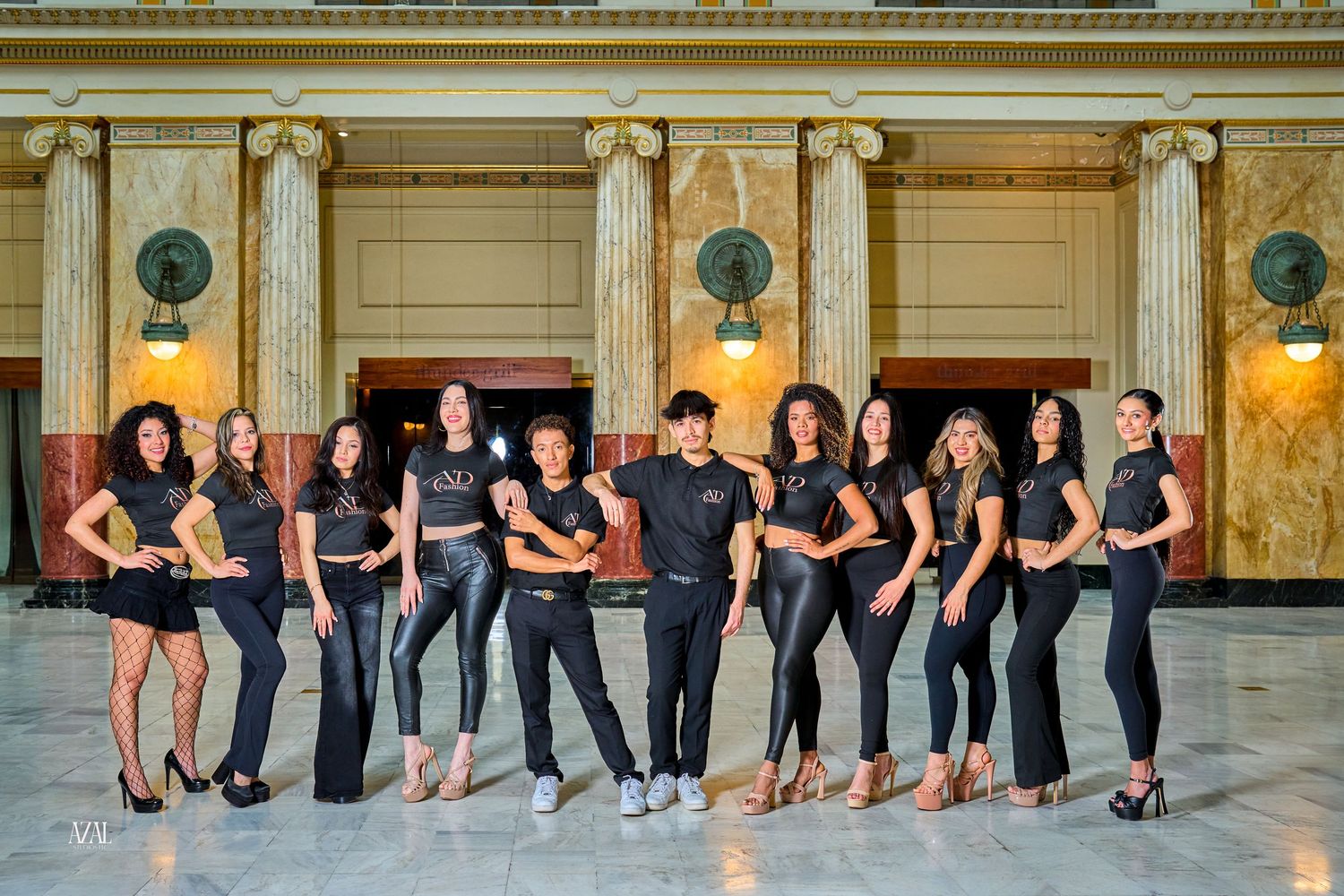 Group of models in matching black outfits pose together in a grand marble-columned ballroom.