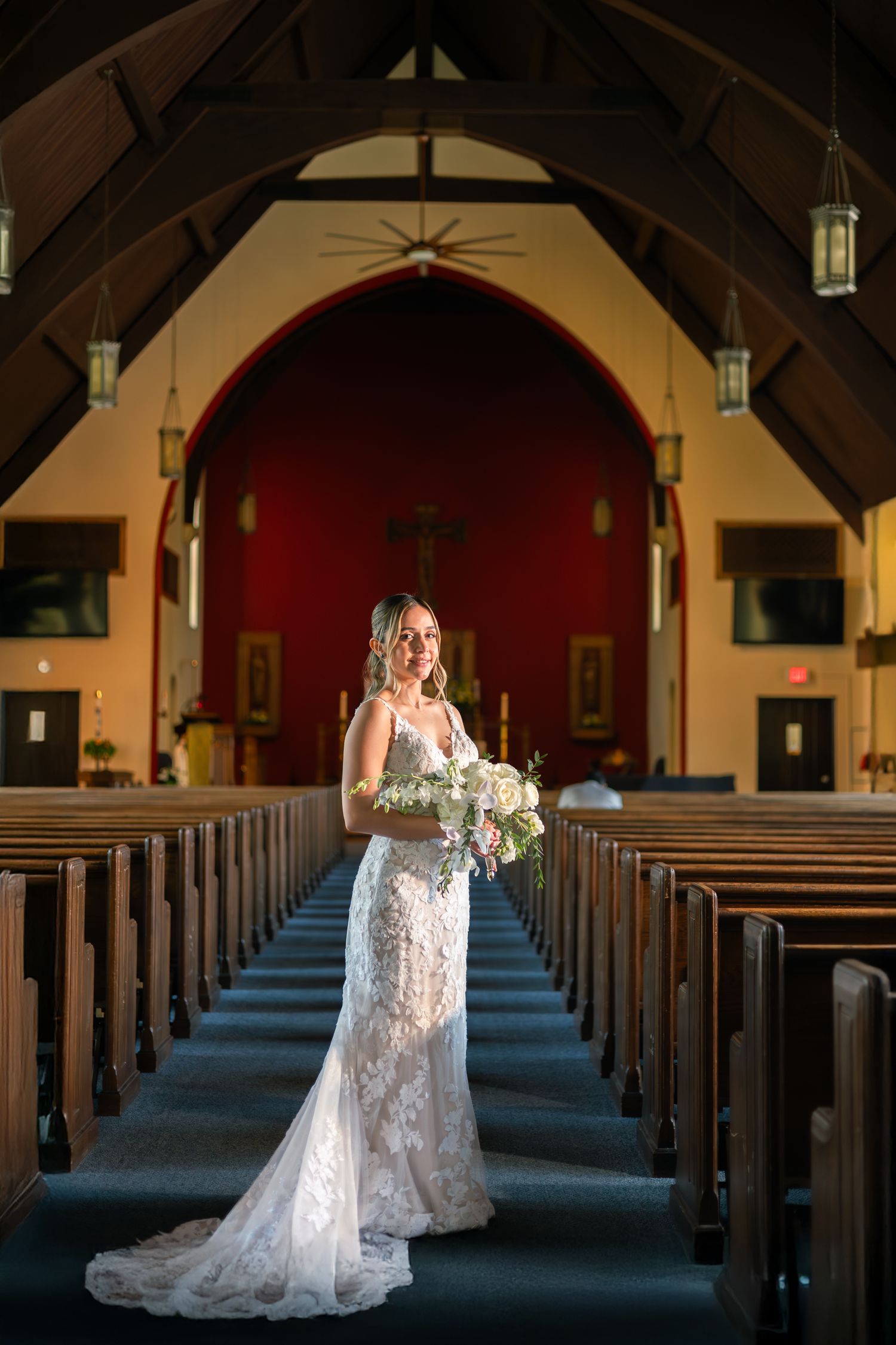A bride in a lace wedding gown stands in the aisle of a church with wooden pews and red walls.