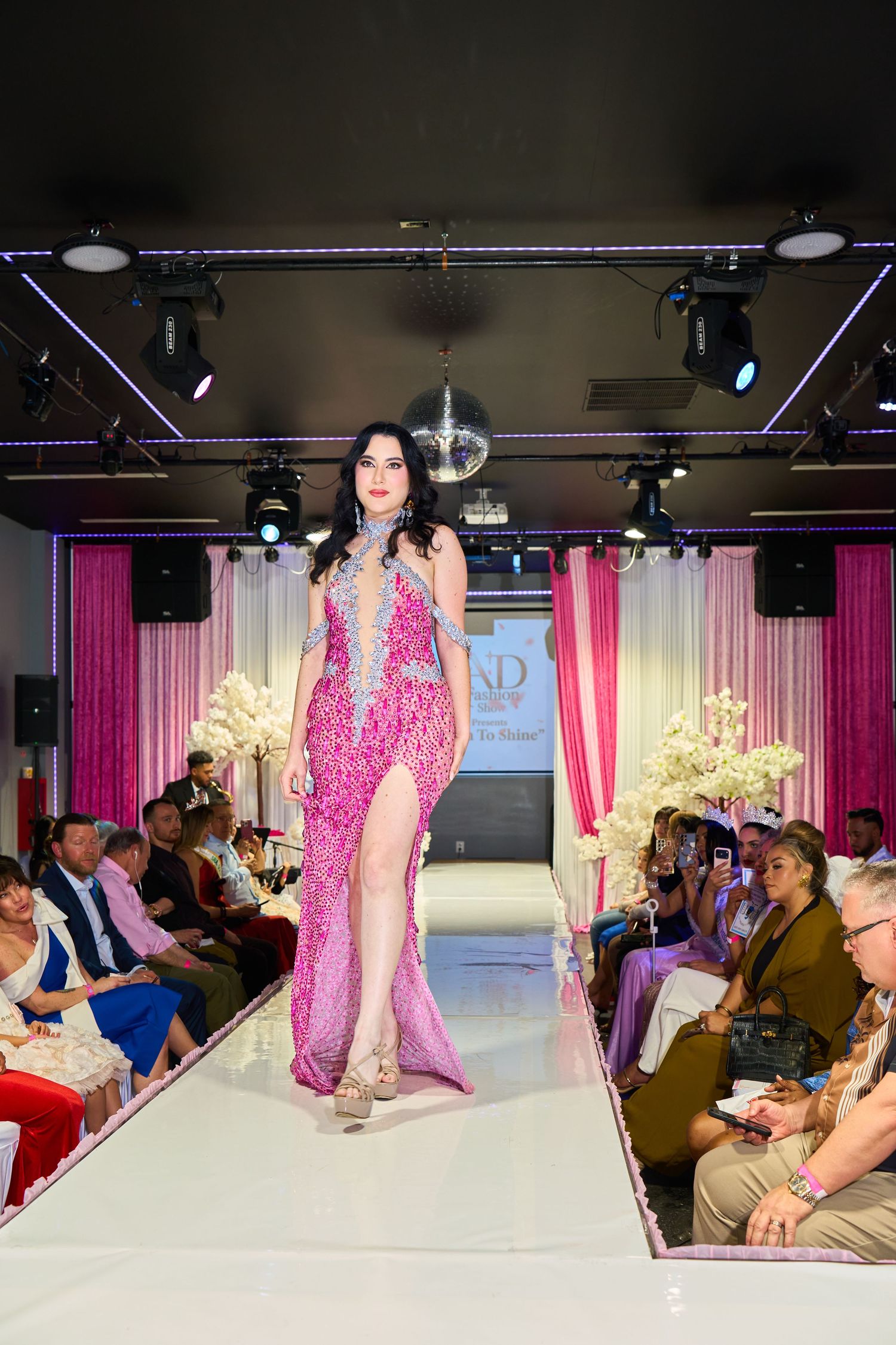 Model showcases a pink sequined gown with high slit on a white runway at a glamorous fashion event.
