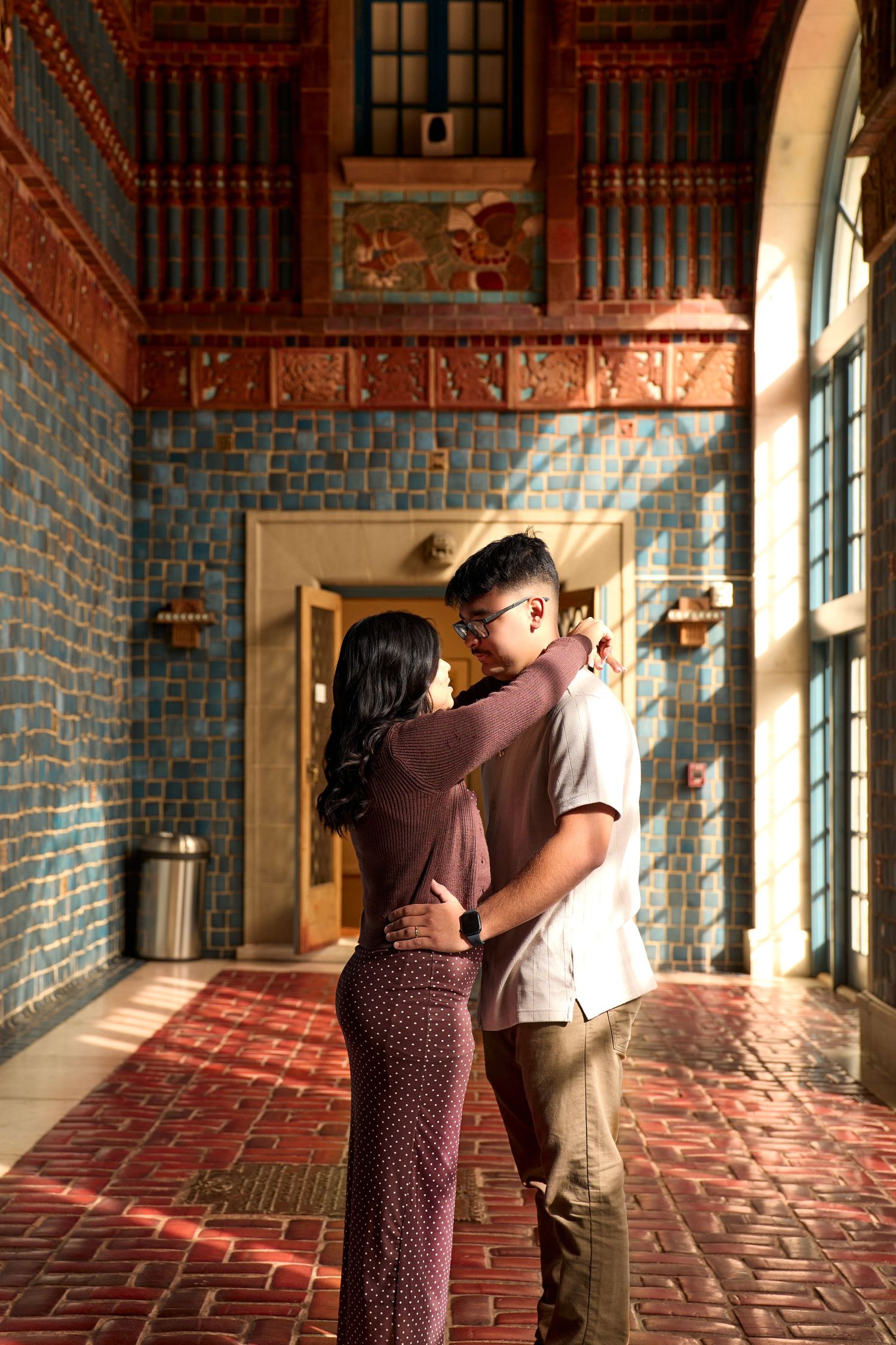A romantic couple embraces in a sunlit brick corridor with arched windows casting warm shadows on the floor.