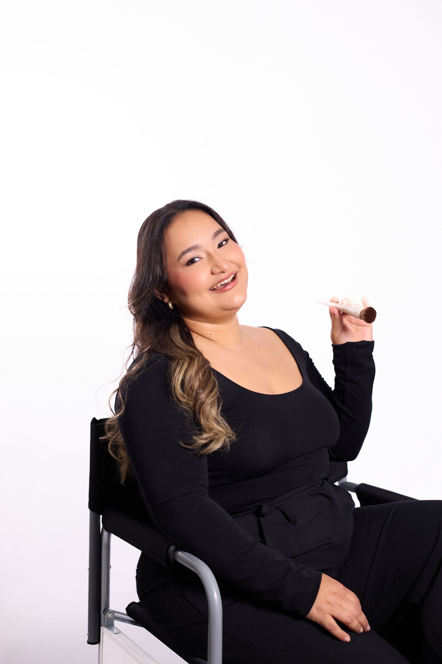 A person in black attire poses playfully in a chair against a white background.