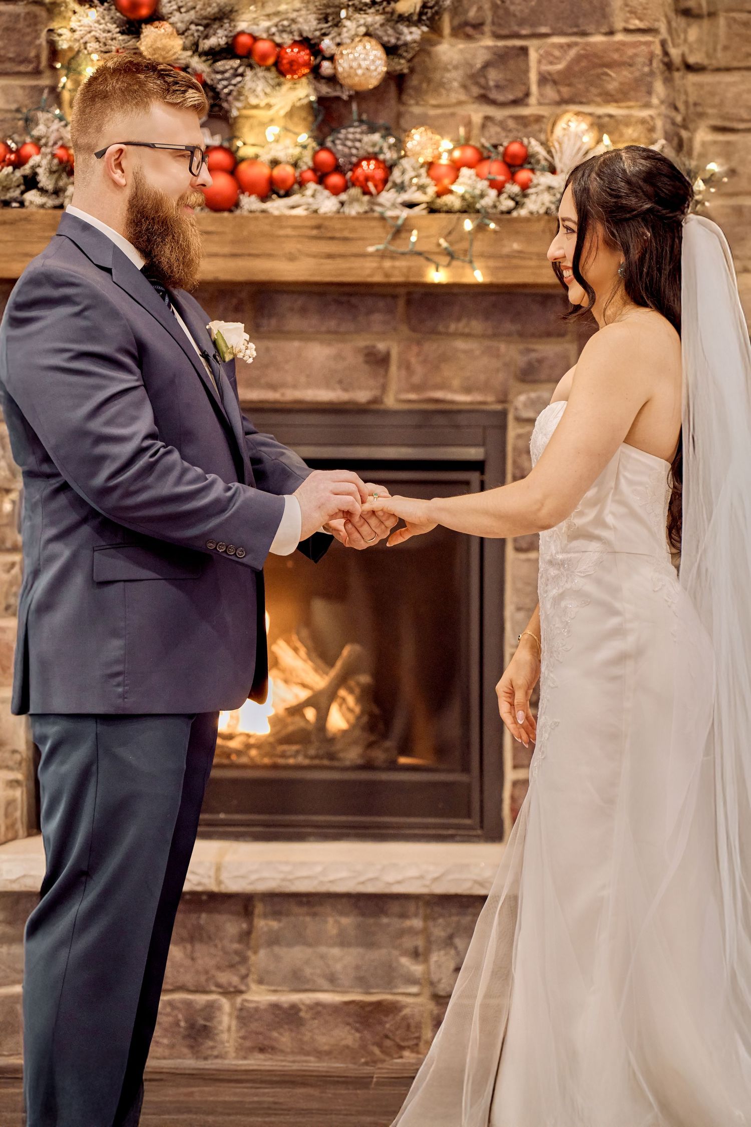 Wedding couple holds hands during intimate ceremony in front of rustic stone fireplace with festive holiday decorations.