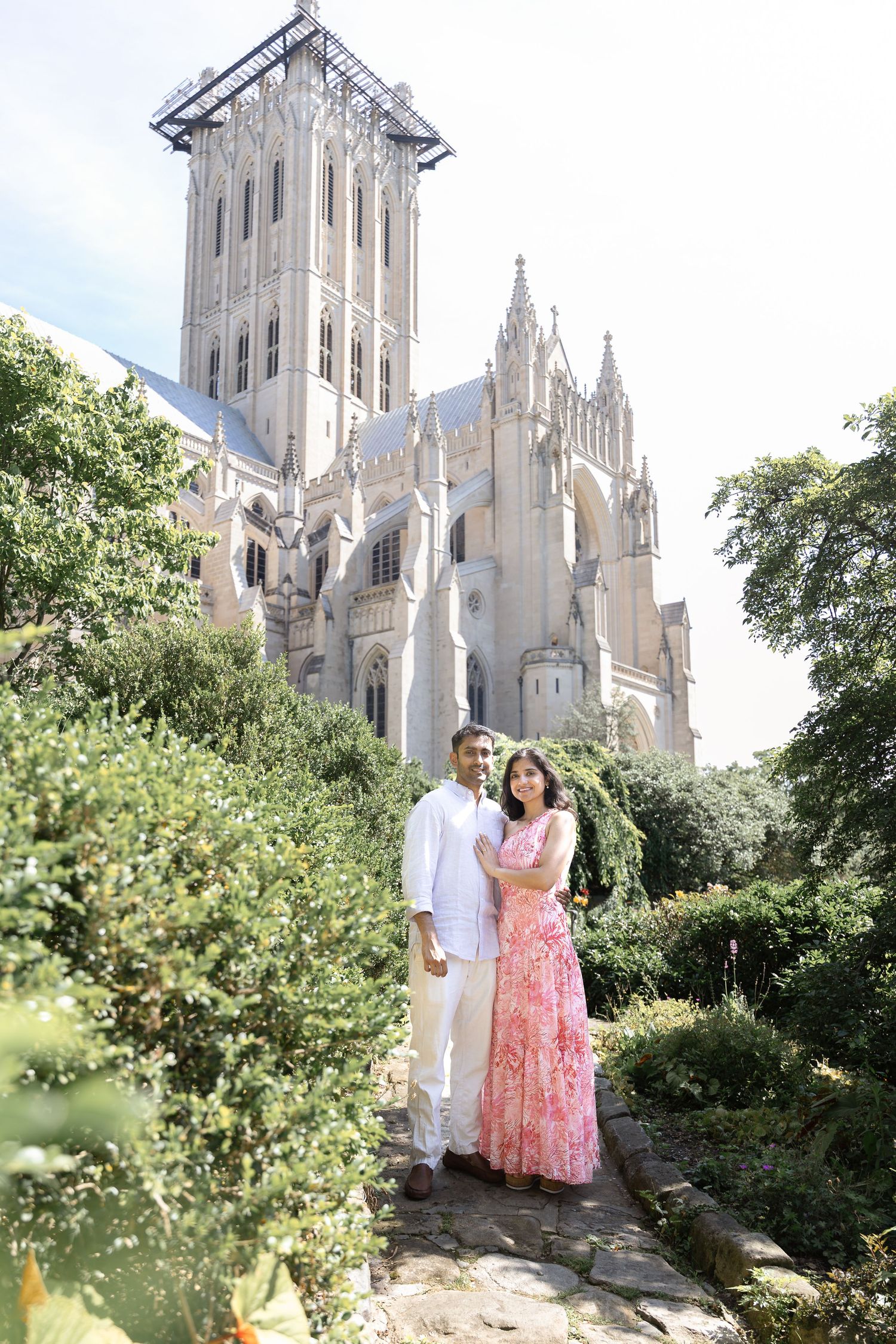 A couple poses against the backdrop of a Gothic cathedral's soaring spires and lush greenery on a sunny day.
