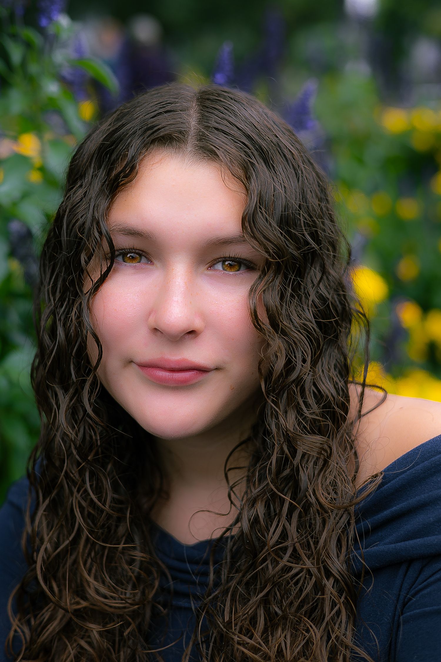 Portrait photo with dark wavy hair in front of blooming purple and yellow flowers in a garden setting.
