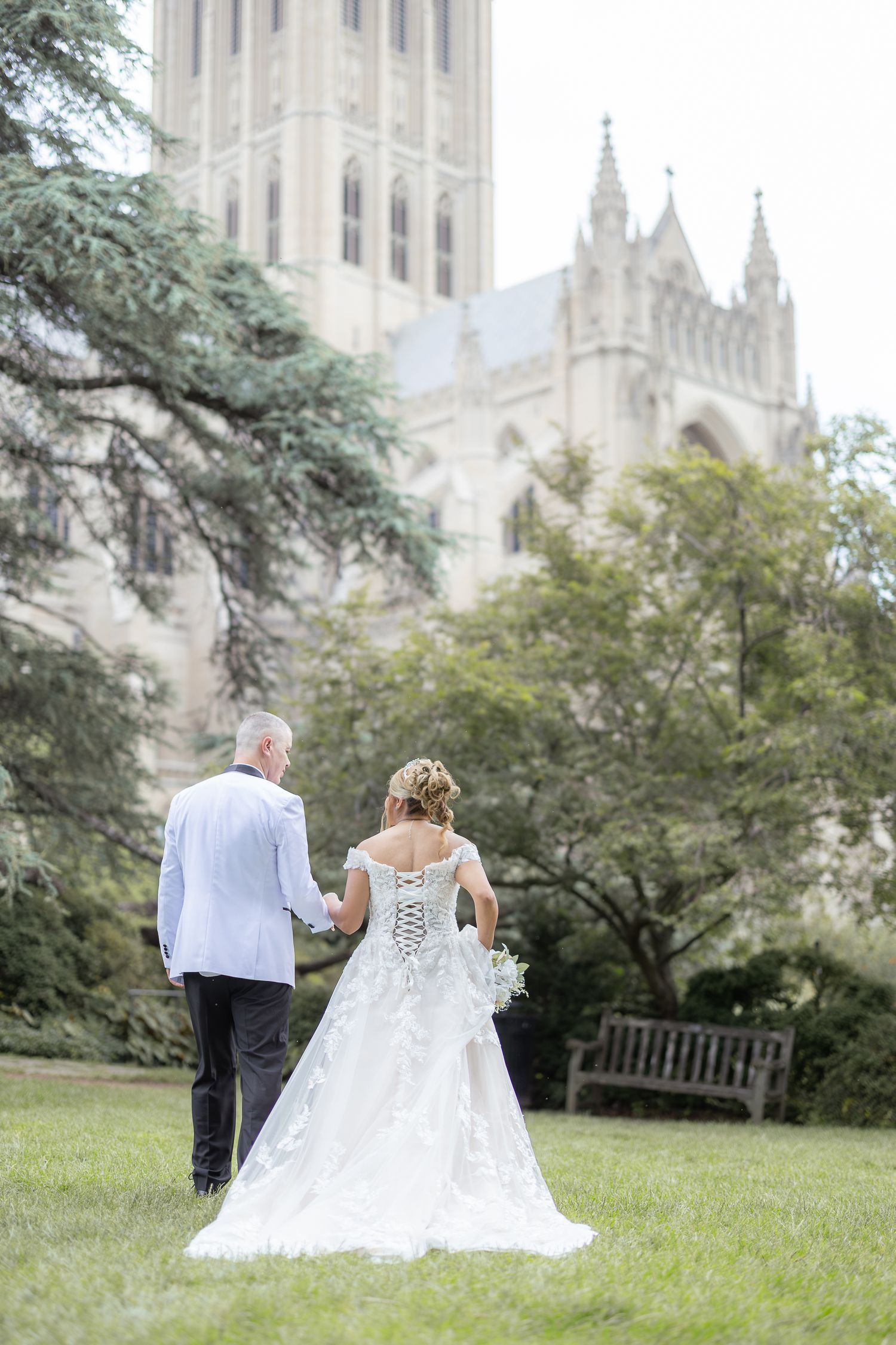 A couple in wedding attire sits together on a wooden bench in front of a grand gothic cathedral on a peaceful summer day.