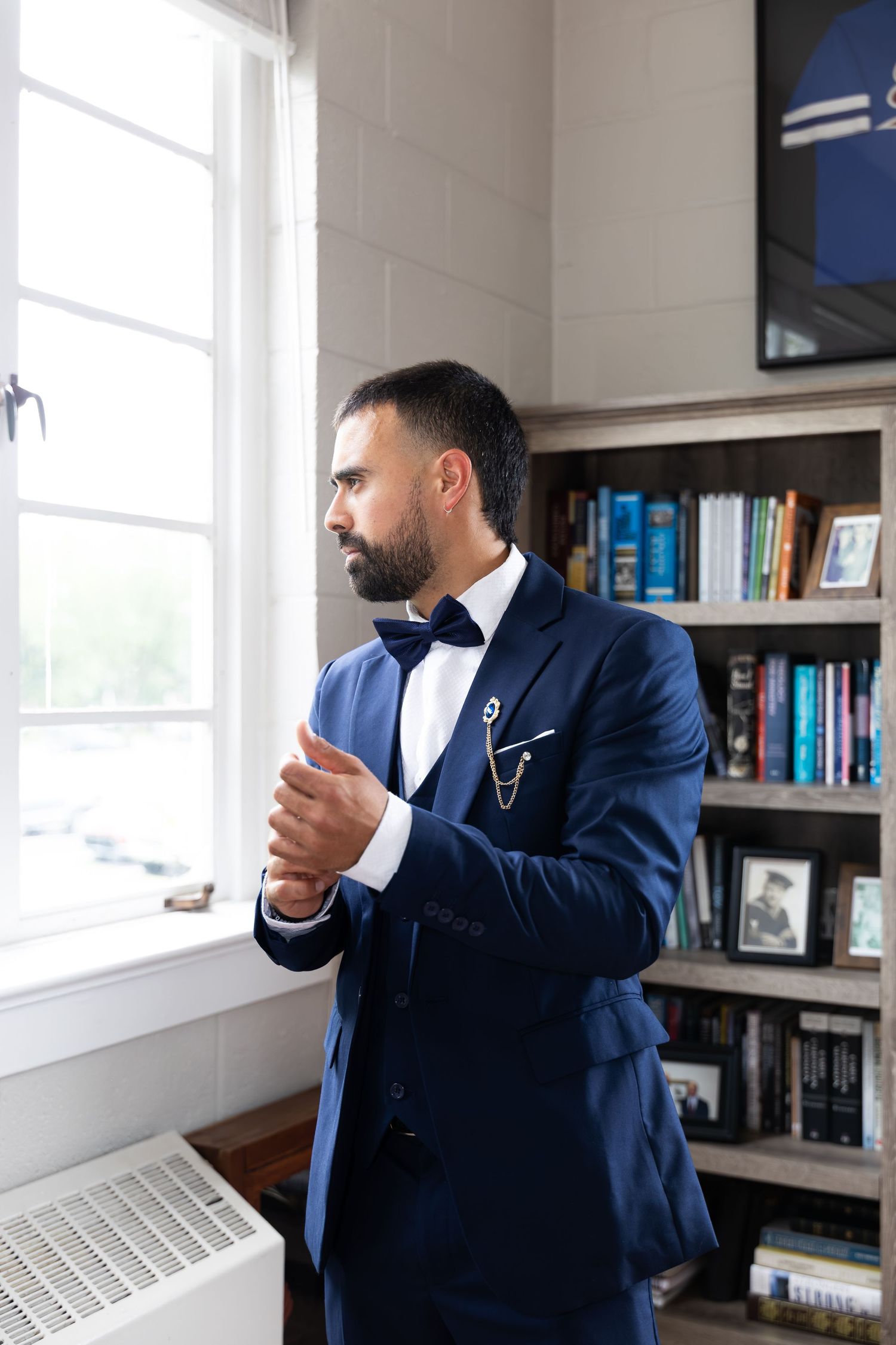 A bearded man in a navy blue tuxedo with bow tie stands by a window next to a bookshelf in an elegant room.
