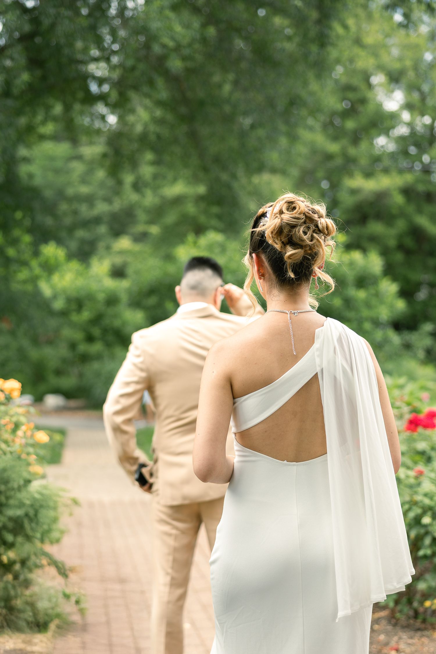 Elegant bride in a one-shoulder white gown with flowing fabric follows groom in tan suit along garden path.