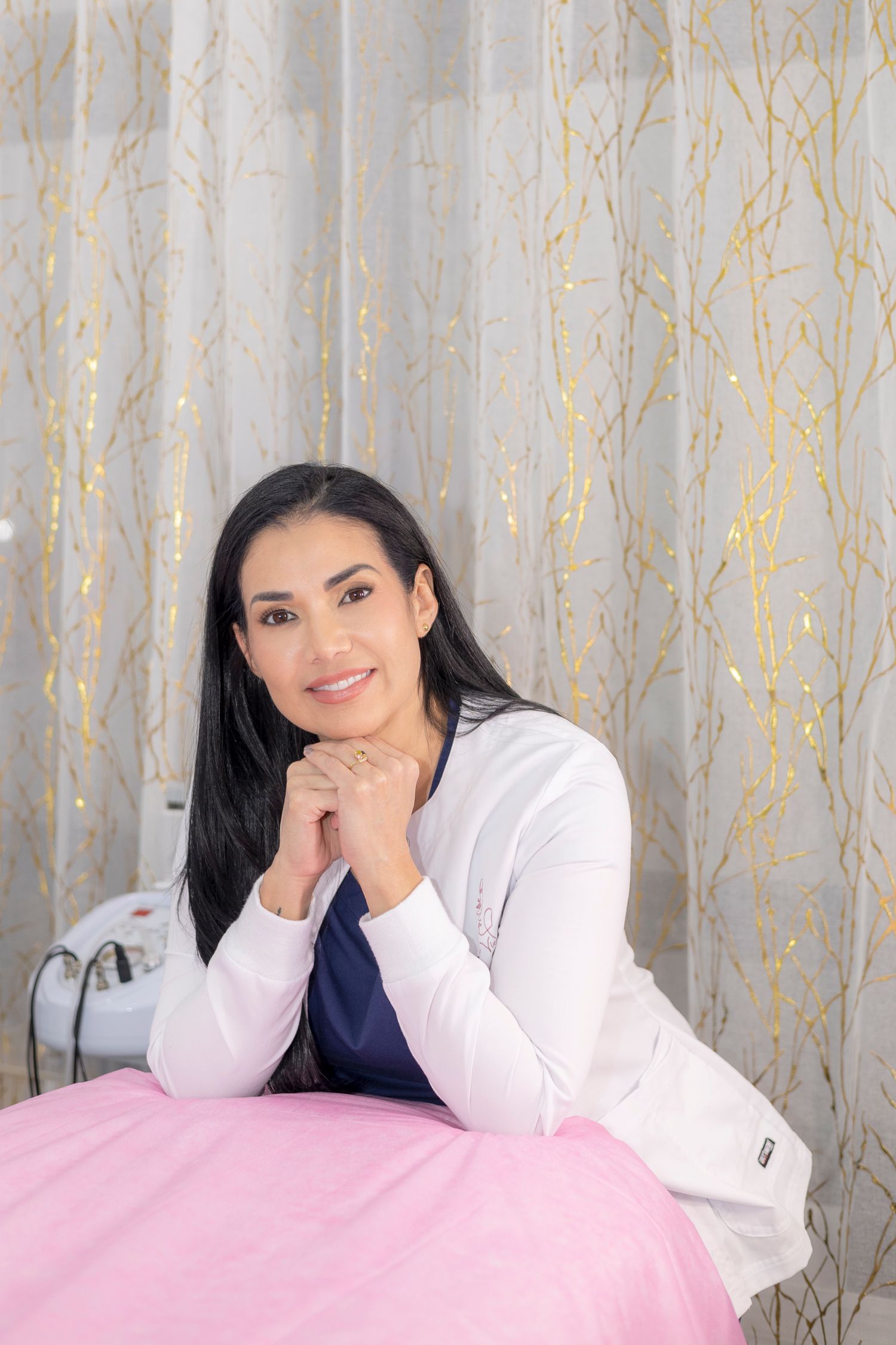 Professional portrait with white cardigan and navy top against silvery curtain background with gold accents.