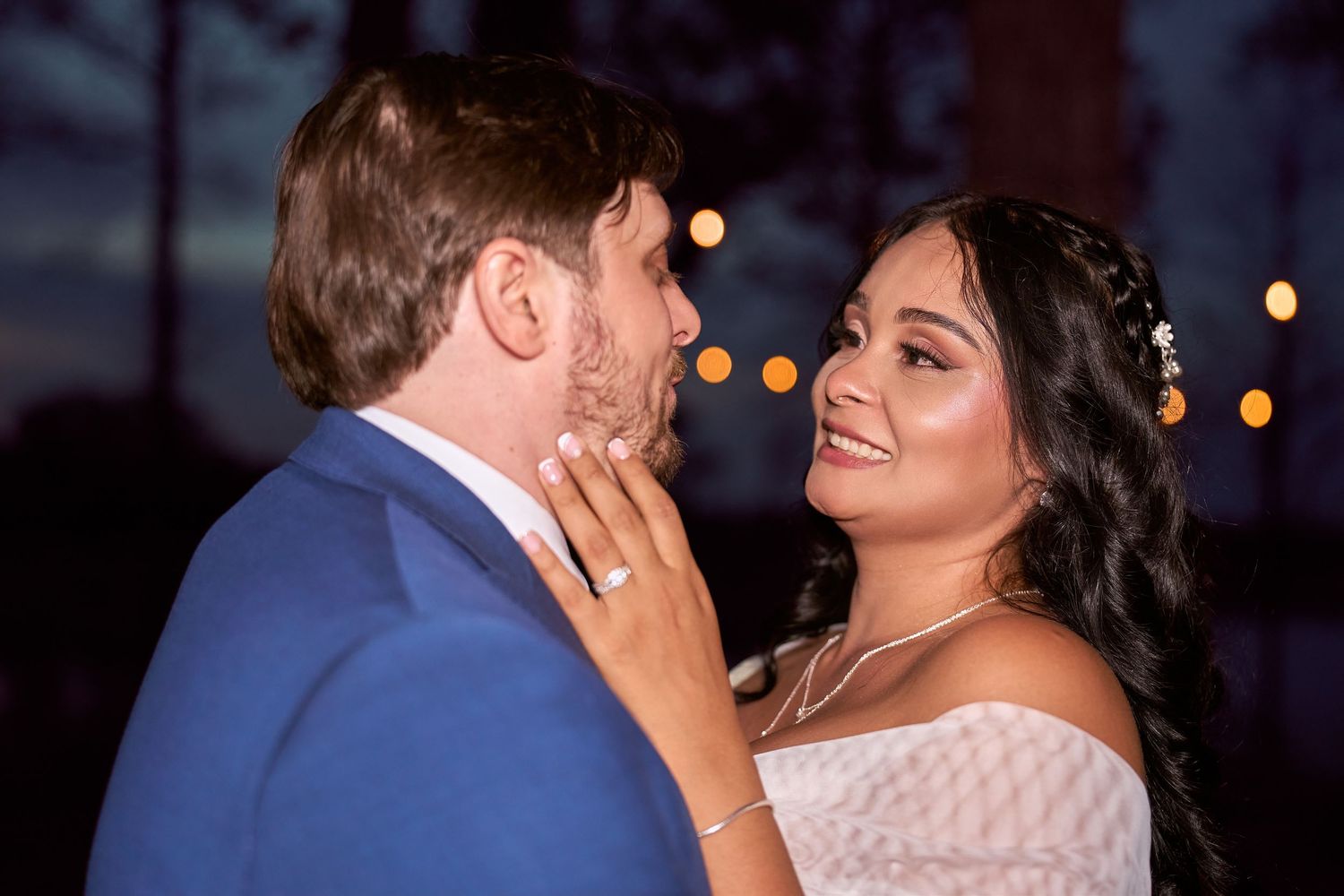 Bride and groom share a romantic moment outdoors at night, with warm bokeh lights glowing in the background.