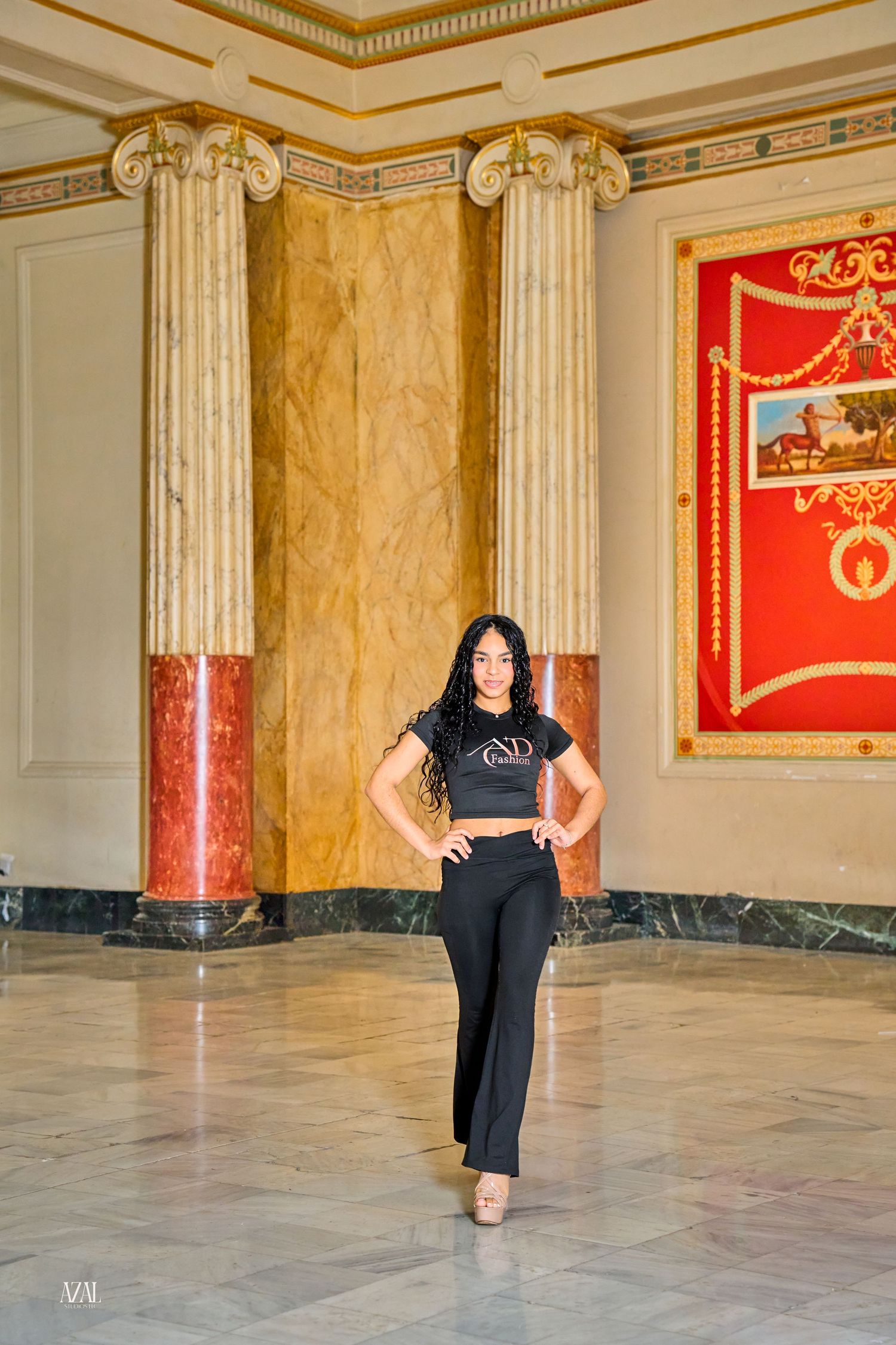Woman in black crop top and flared pants poses confidently in elegant marble hall with ornate columns.