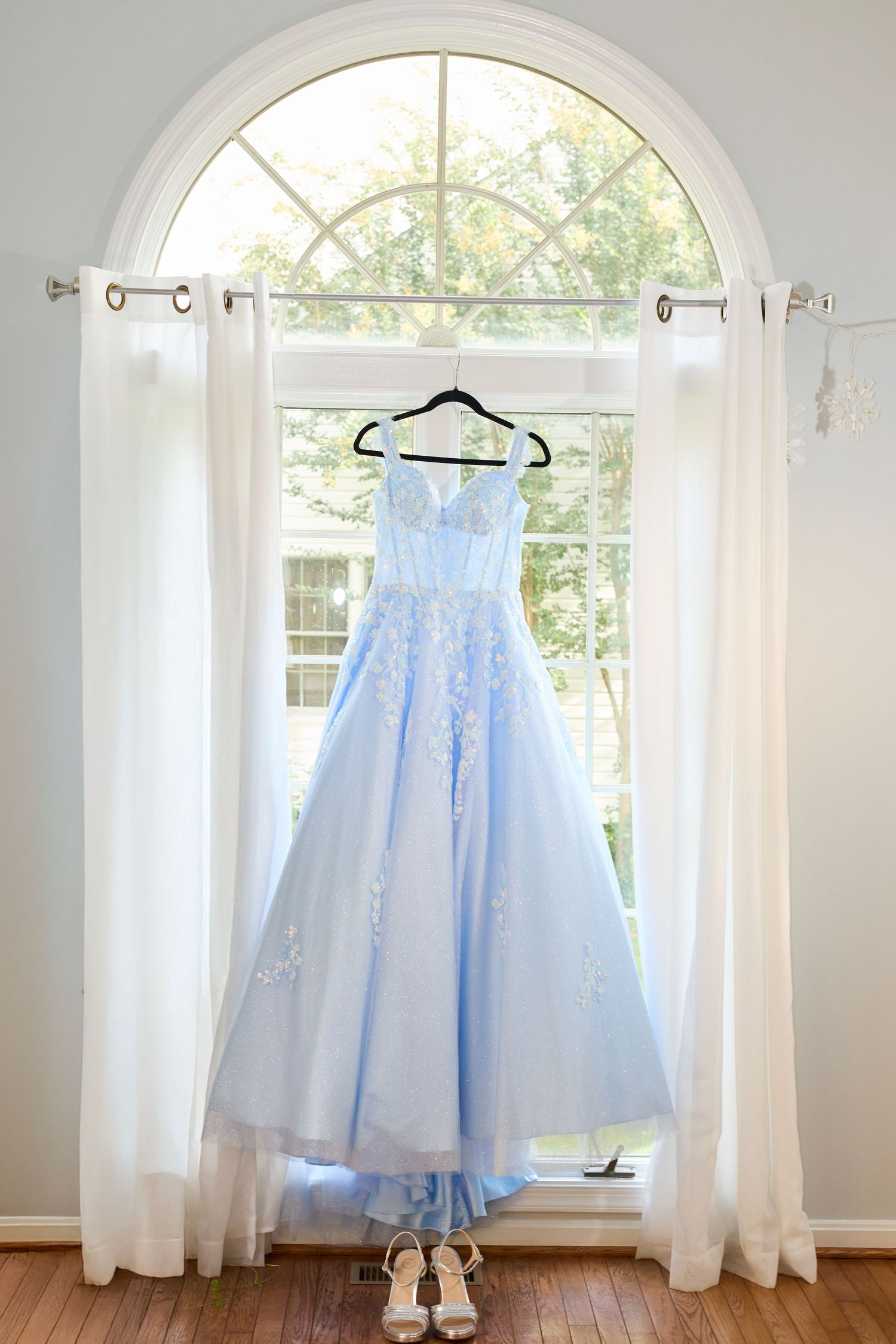 Light blue formal gown with lace details hangs in front of arched window with white curtains and matching shoes below.