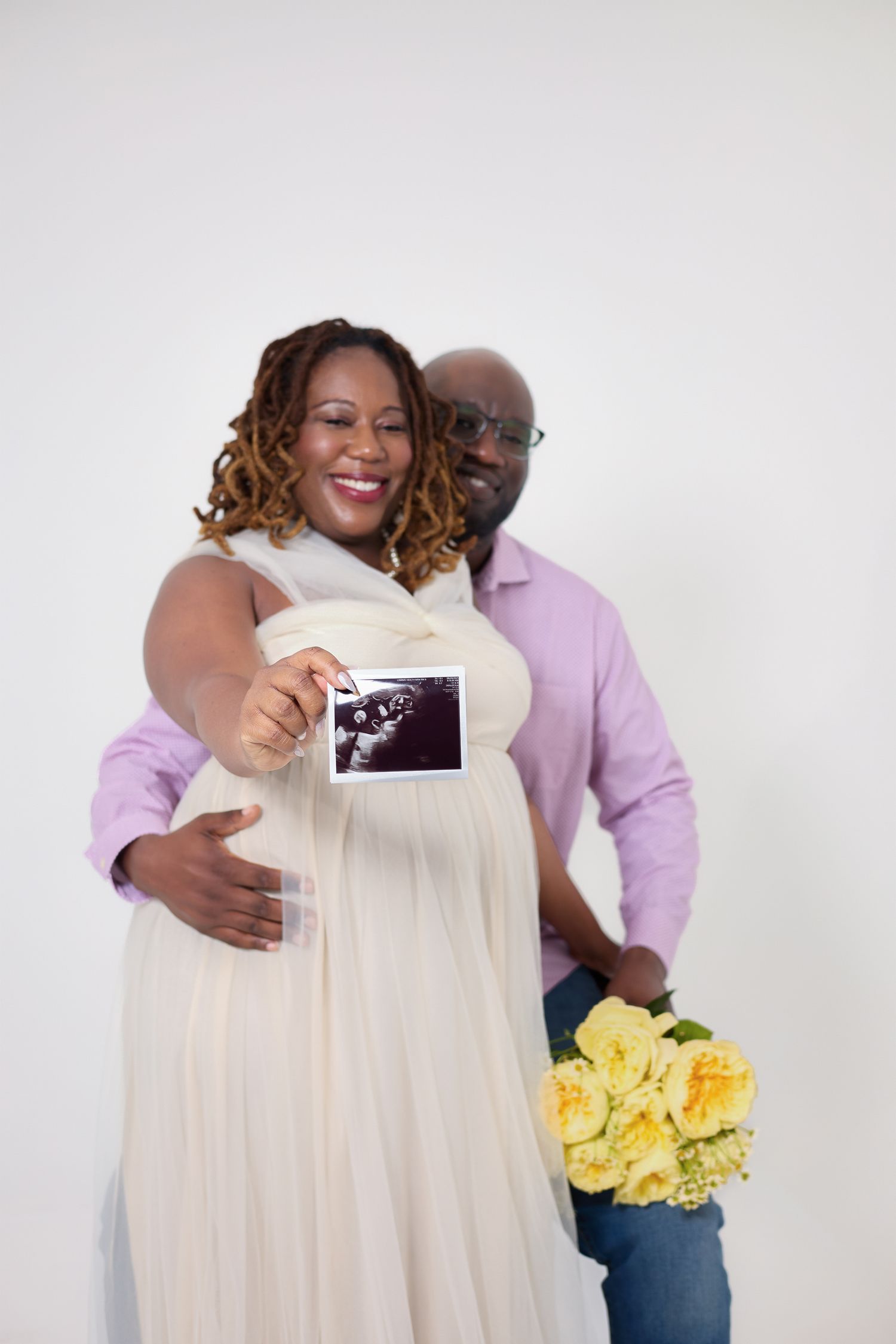 Smiling pregnant Black couple holding ultrasound photo and yellow roses, woman in cream dress, man in pink shirt.