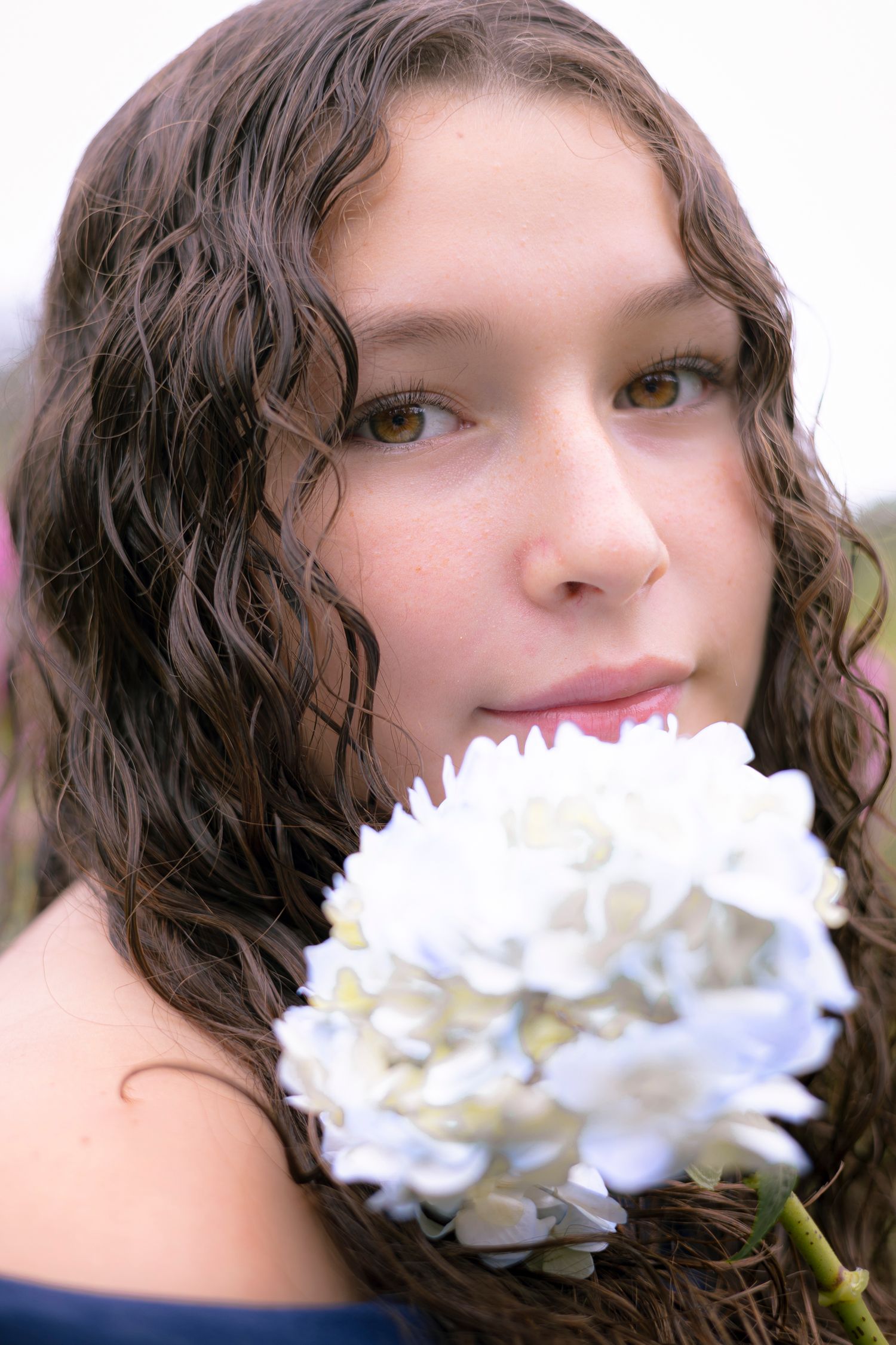 Person with dark curly hair smiles while holding a white carnation flower against a blurred background.