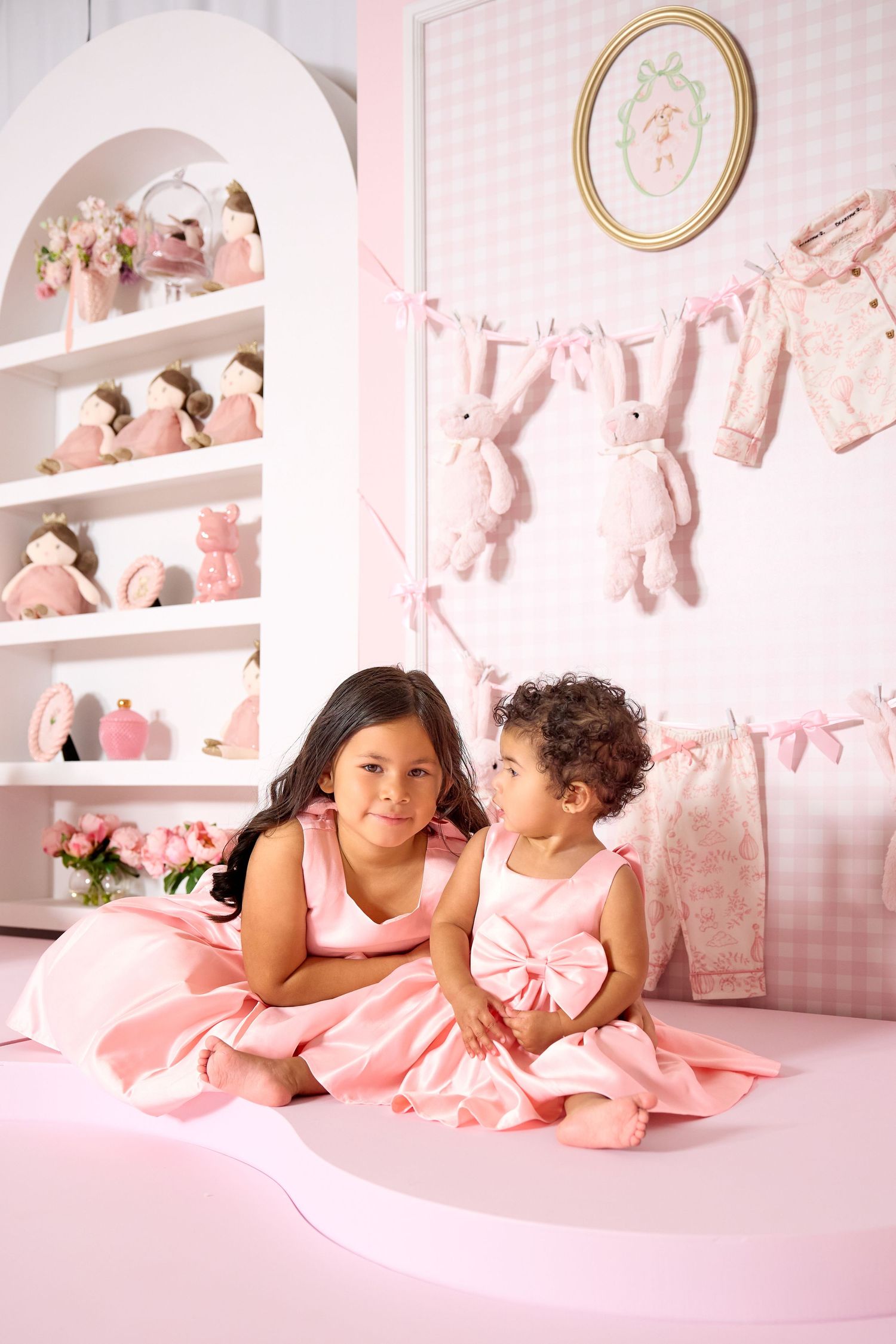 Two young girls in matching pink dresses sit together in a charming pink nursery room with bunny decorations.