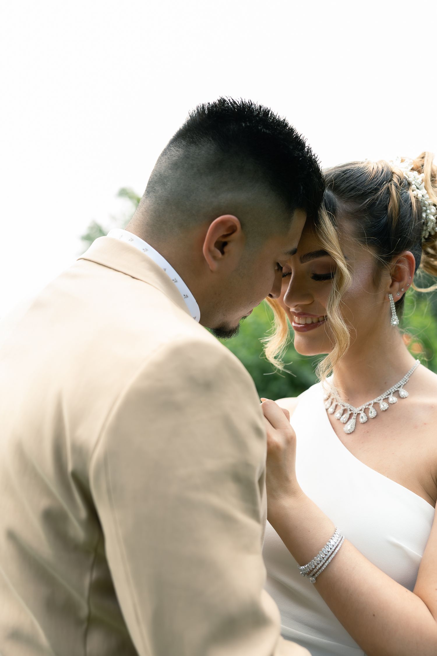 Intimate wedding couple sharing tender moment outdoors with bride wearing elegant necklace and groom in tan suit.