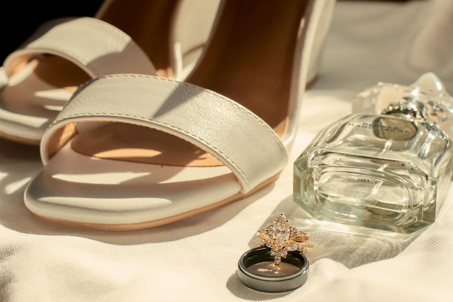A pair of beige leather sandals with a minimal design displayed next to a clear glass perfume bottle on a light surface.