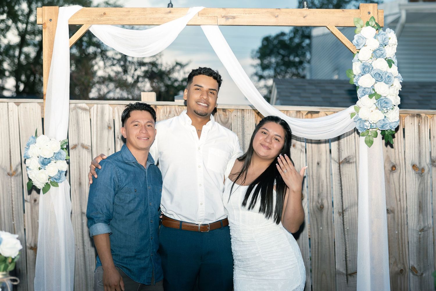 Joyful group poses together at outdoor wedding celebration with white floral decorations and wooden fence backdrop.