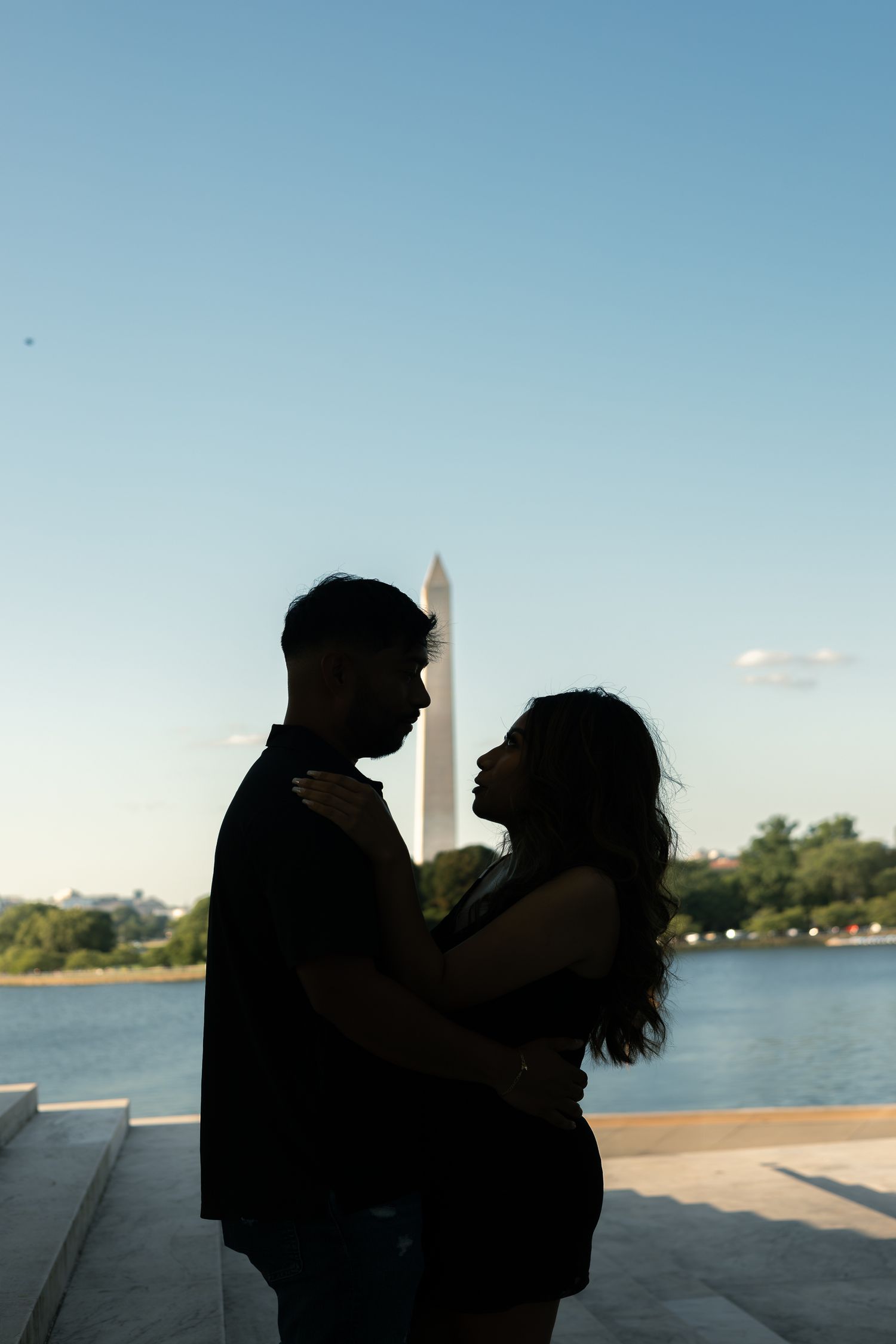 Silhouetted figures embrace on a waterfront promenade with a blue sky and water in the background.