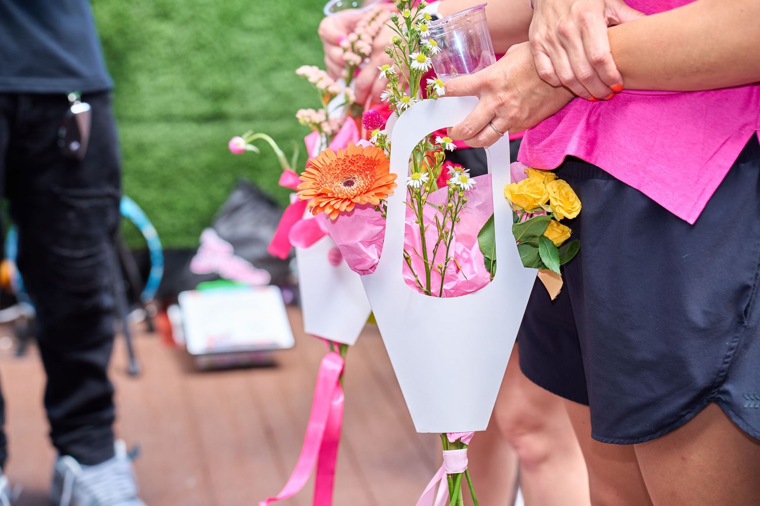 People holding colorful flower bouquets wrapped in white paper with pink ribbons at an outdoor event.